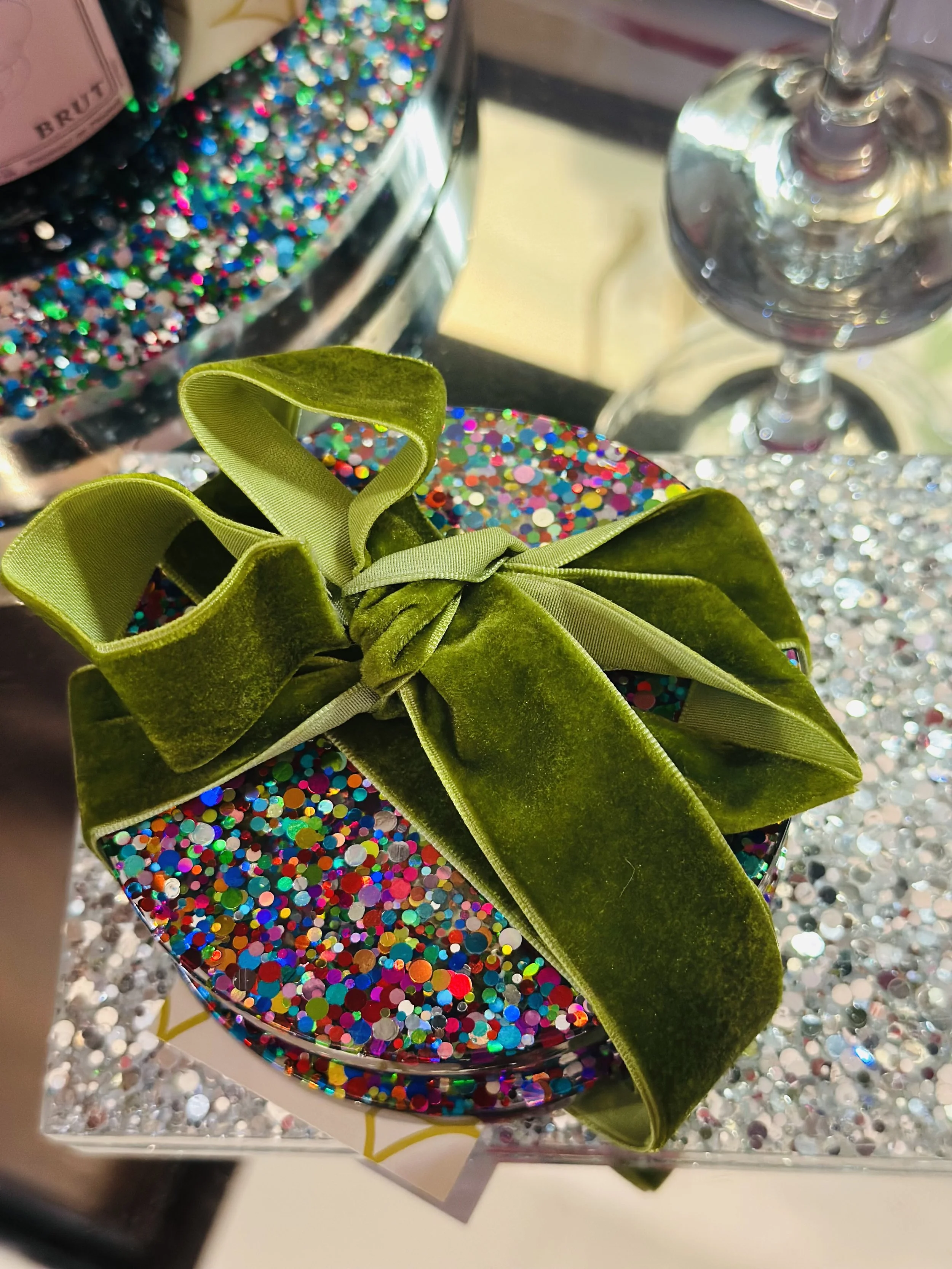 Colorful gift box decorated with multicolored glitter, topped with a green velvet ribbon bow, set on a glittery surface with a background of additional glitter and a wine glass.