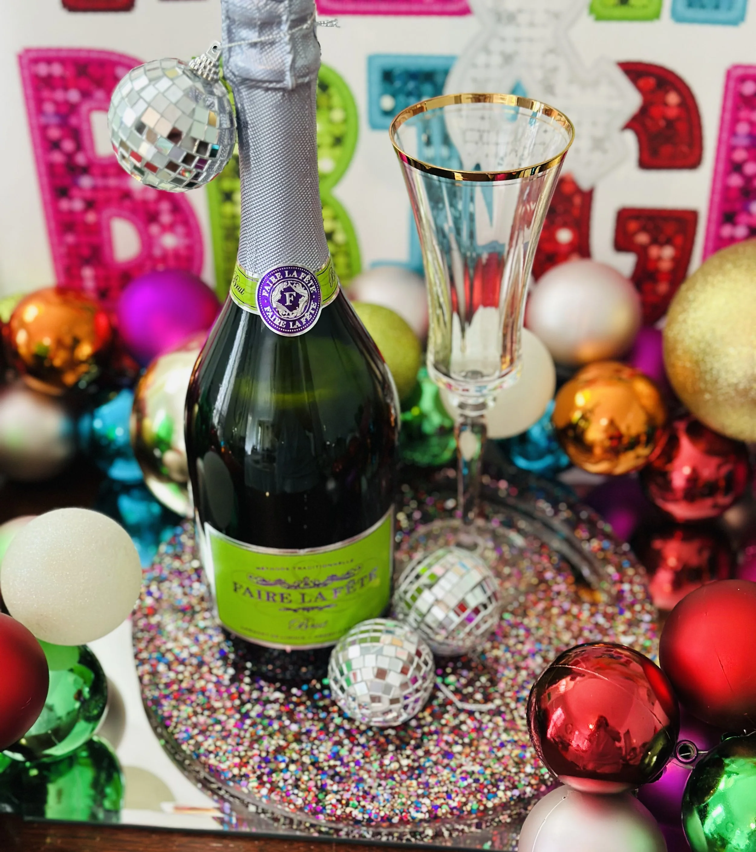 A bottle of sparkling wine with a disco ball ornament attached to its neck, placed on a glittery surface surrounded by colorful Christmas ornaments and decorations, with a festive backdrop.