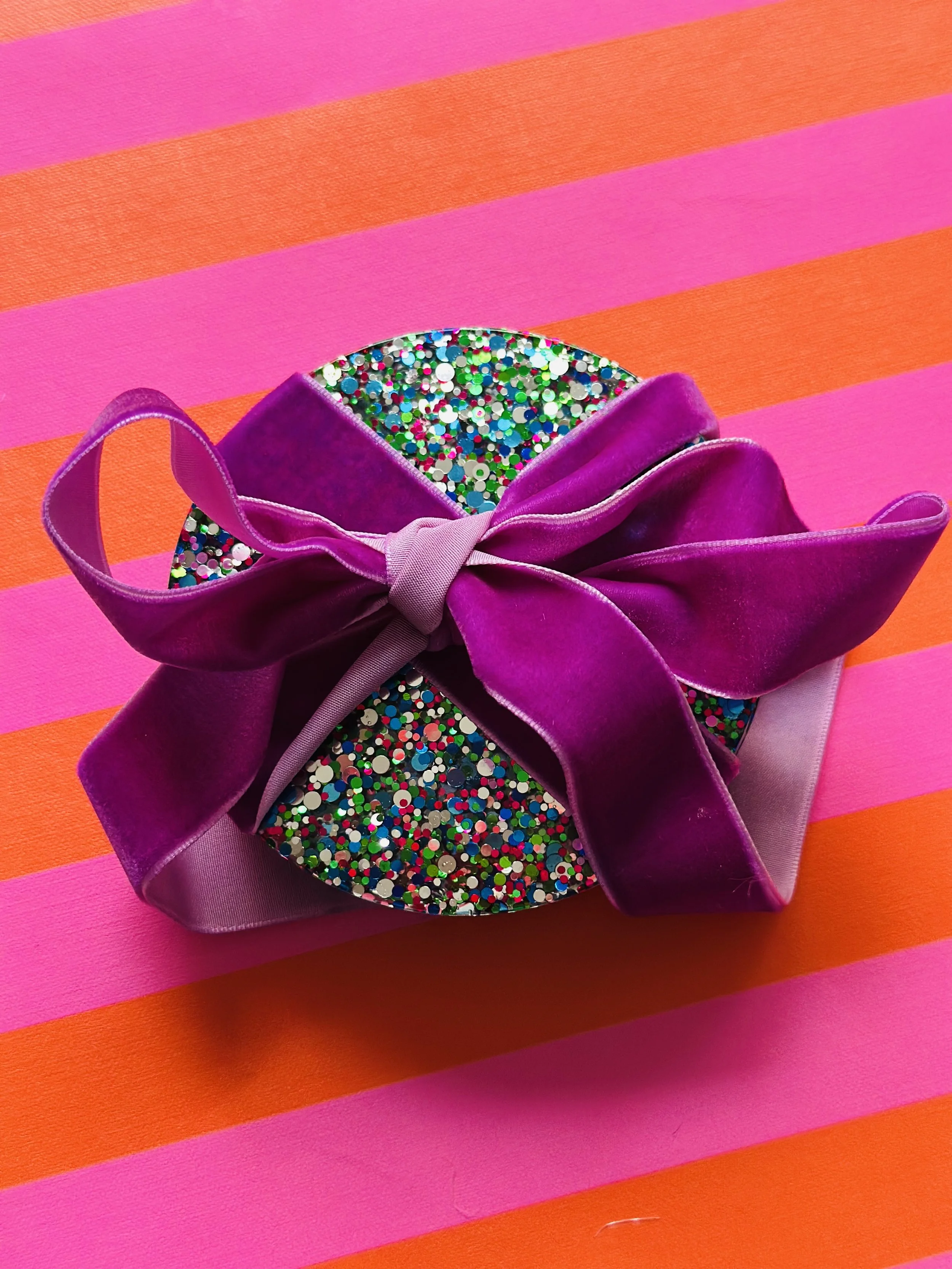Colorful gift box decorated with multicolored glitter and a purple satin ribbon bow, placed on pink and orange striped background.