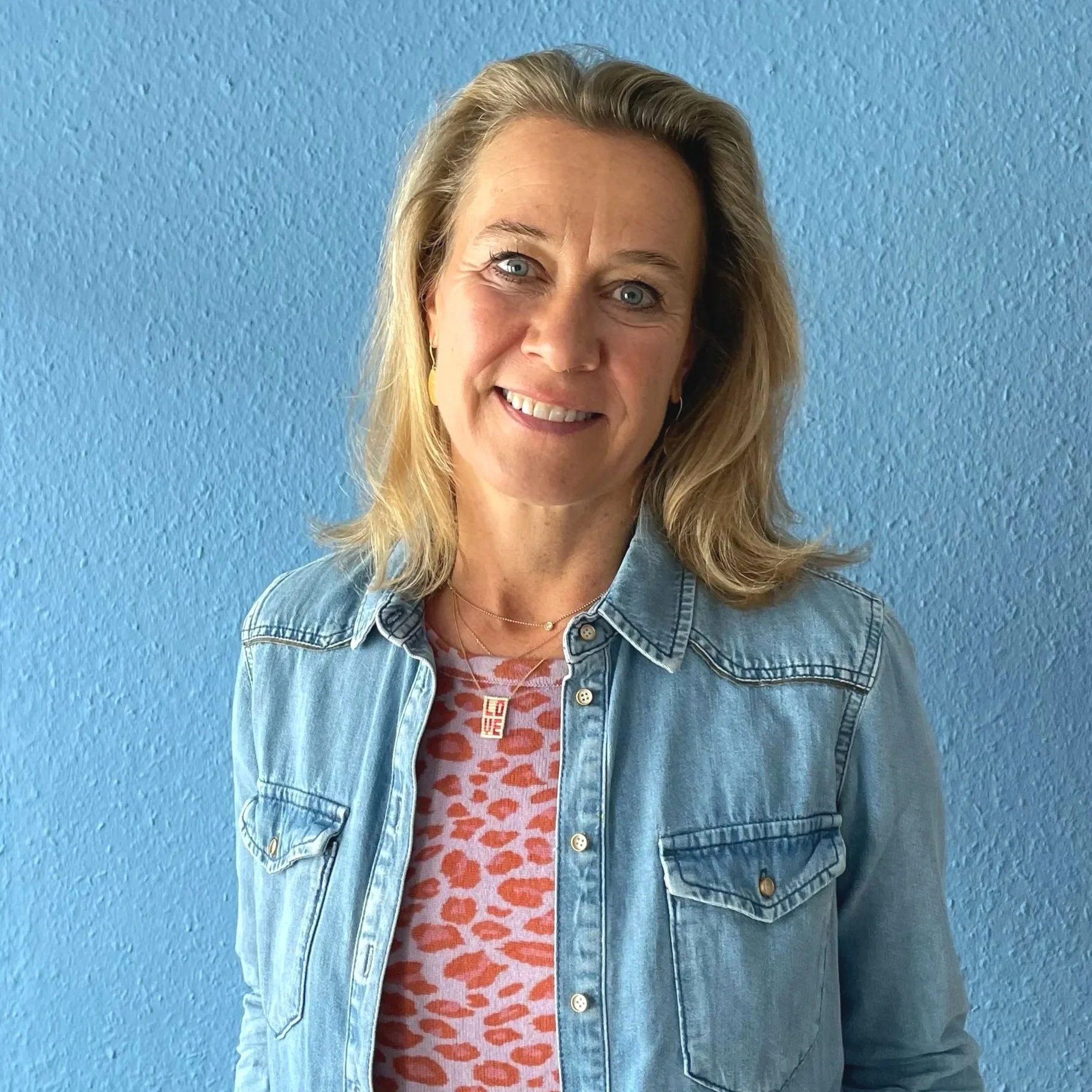 Eine lächelnde Frau mit blonden, schulterlangen Haaren vor einer blauen Wand, trägt eine Denimjacke und ein rotes Oberteil mit Leopardenmuster.
