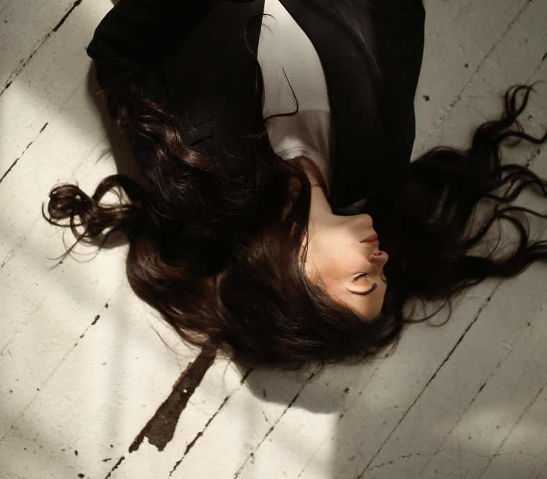 A woman with long dark hair lying on a white wooden floor, eyes closed, wearing a black jacket and white shirt.