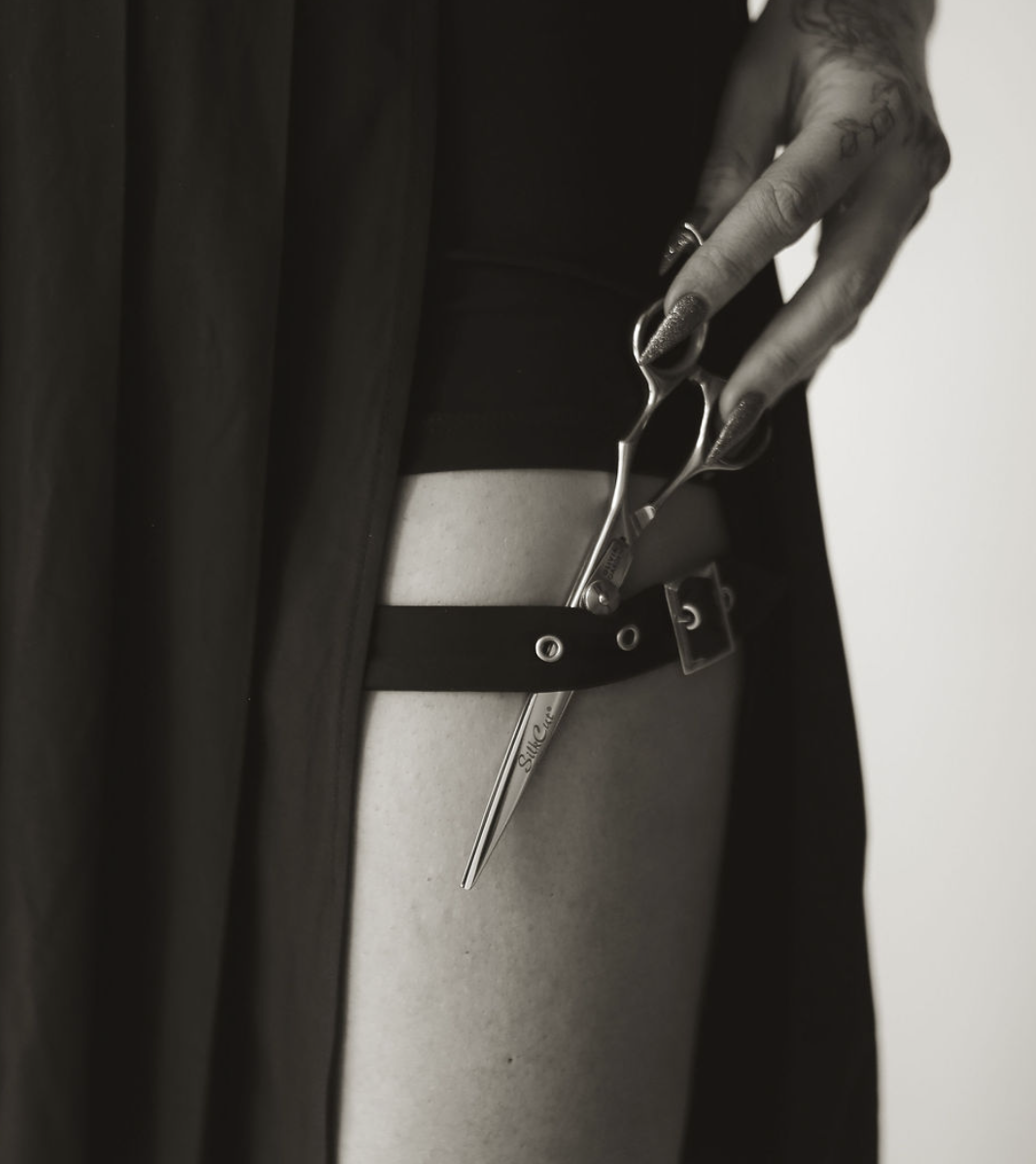 A tattooed person in dark clothing using surgical scissors to cut a strap around their thigh with a tattoo on their thigh.