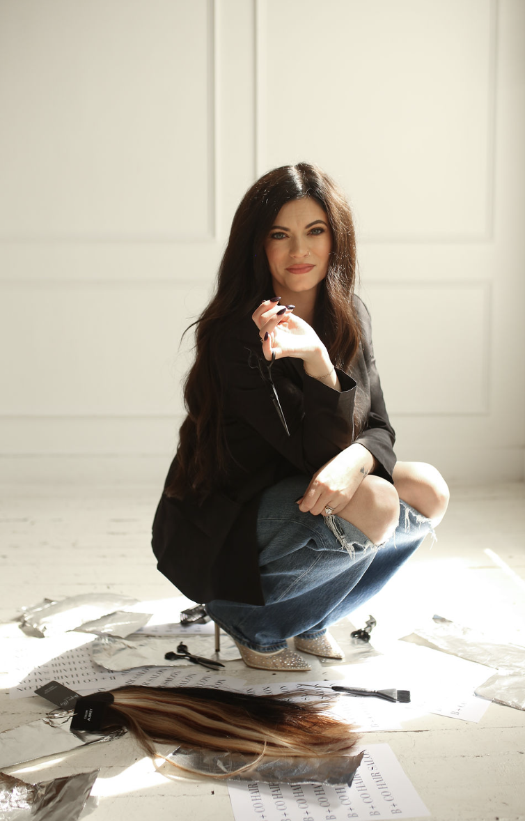 A woman crouching on a white floor surrounded by torn paper and foil, with hair extensions in front of her and a neutral background.