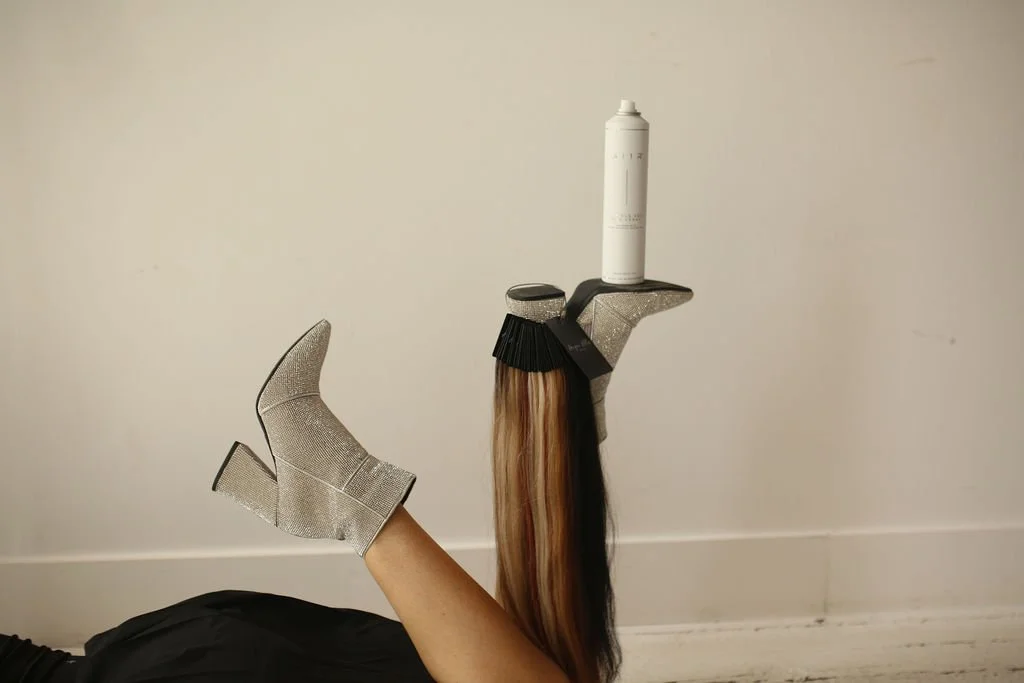 A hairdressing mannequin arm wearing a silver high heel shoe, with a hairbrush and a bottle of hair product balanced on top of the heel, all against a plain white wall.