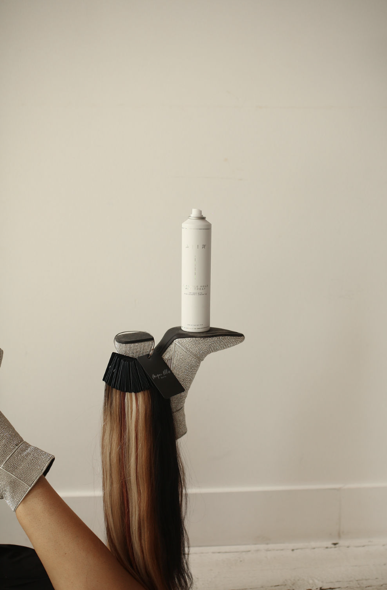 A pair of gloves, a black hairbrush, and a hair extension with blonde and black hair are balanced on a hand holding a hair styling tool. A white aerosol can is placed on the tool.