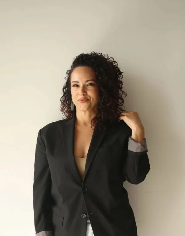 A woman with curly dark hair smiling, wearing a black blazer and light-colored pants, sitting against a plain white wall