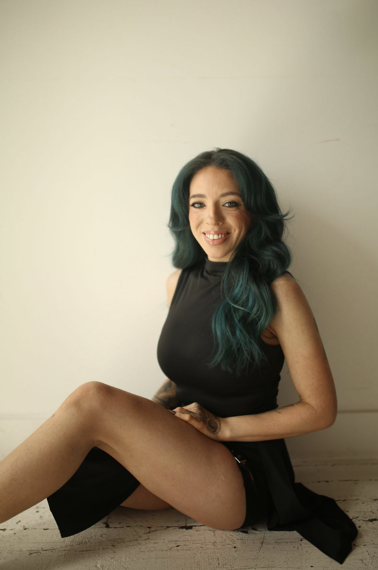 A woman with teal hair sitting on the floor, smiling, wearing a black sleeveless top and shorts, with a tattoo on her left arm, against a plain light-colored background.