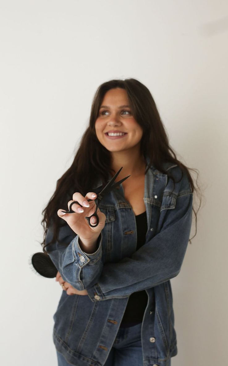 A smiling woman with long dark hair holding a pair of scissors, standing against a plain light-colored background.