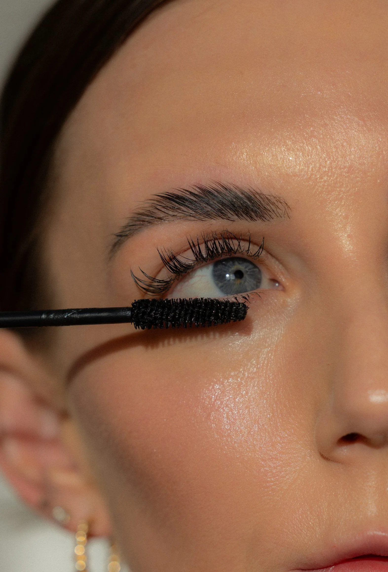 Close-up of a woman applying black mascara to her upper eyelashes using a mascara wand. She has fair skin, blue eyes, neatly groomed eyebrows, and is wearing gold earrings.