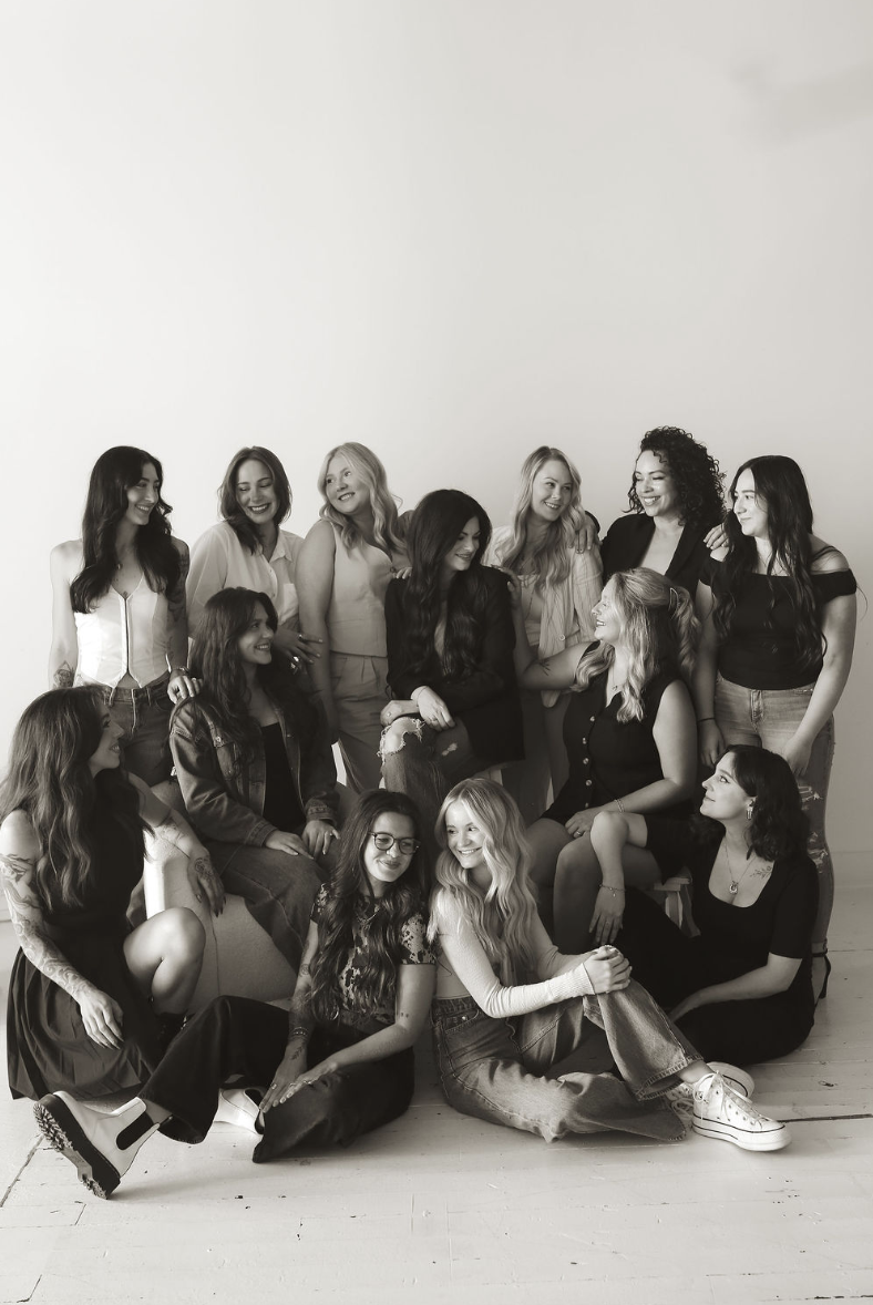 A group of 13 women, some sitting, some standing, smiling and interacting, in a photo shoot against a plain white wall, black and white image.