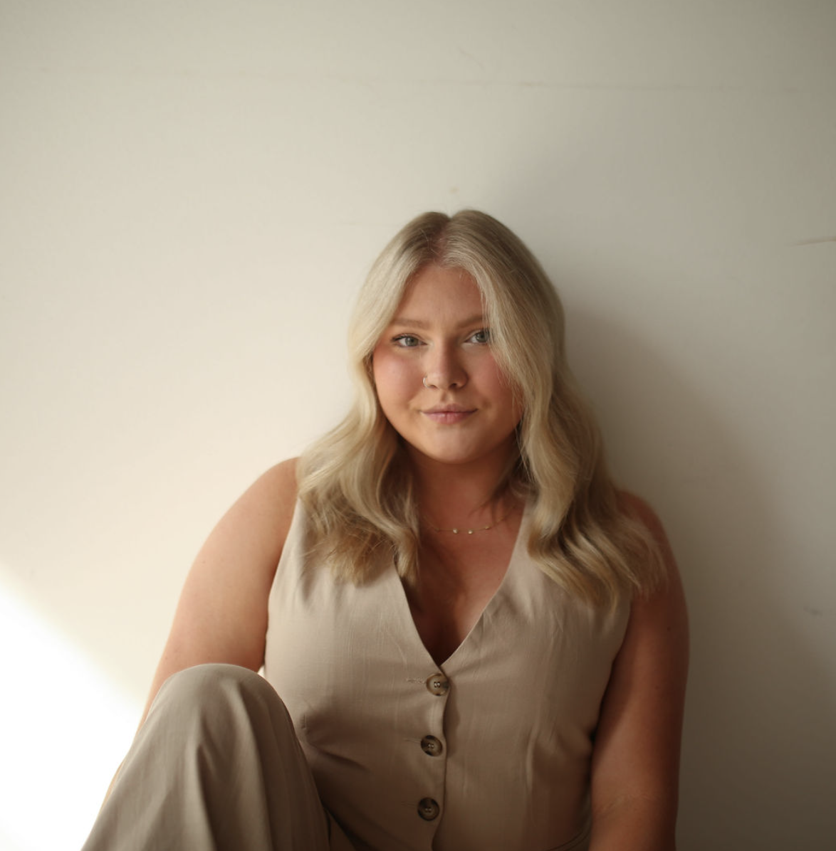A young woman with blonde hair, blue eyes, and a nose piercing, sitting against a plain white wall, wearing a sleeveless beige top with buttons and a gold necklace.