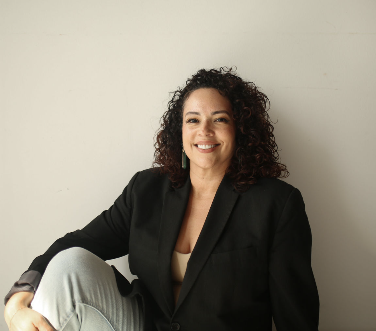A woman with curly dark hair smiling, wearing a black blazer and light-colored pants, sitting against a plain white wall