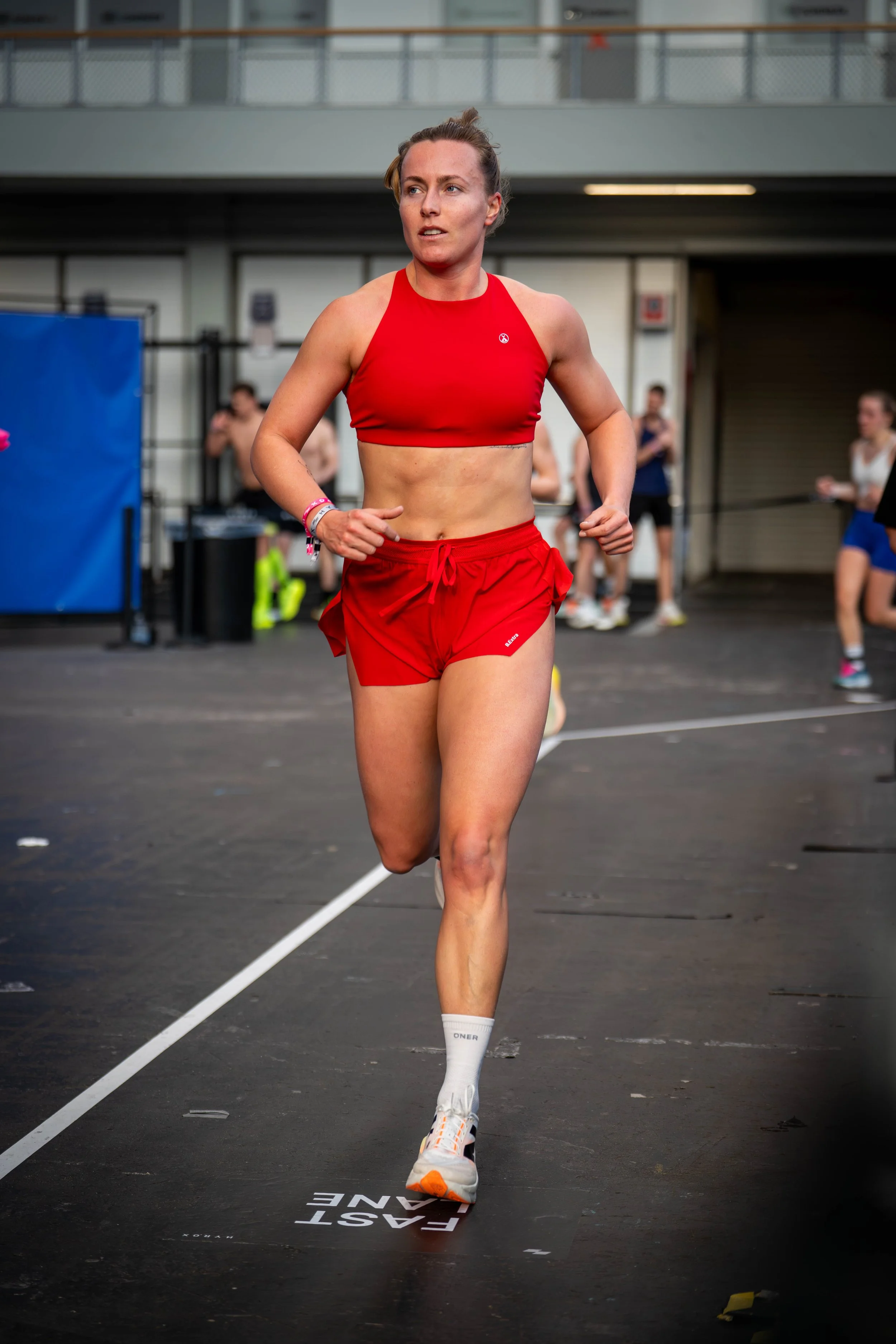 A woman in a red sports bra and red shorts running indoors on a track, with other people in the background.