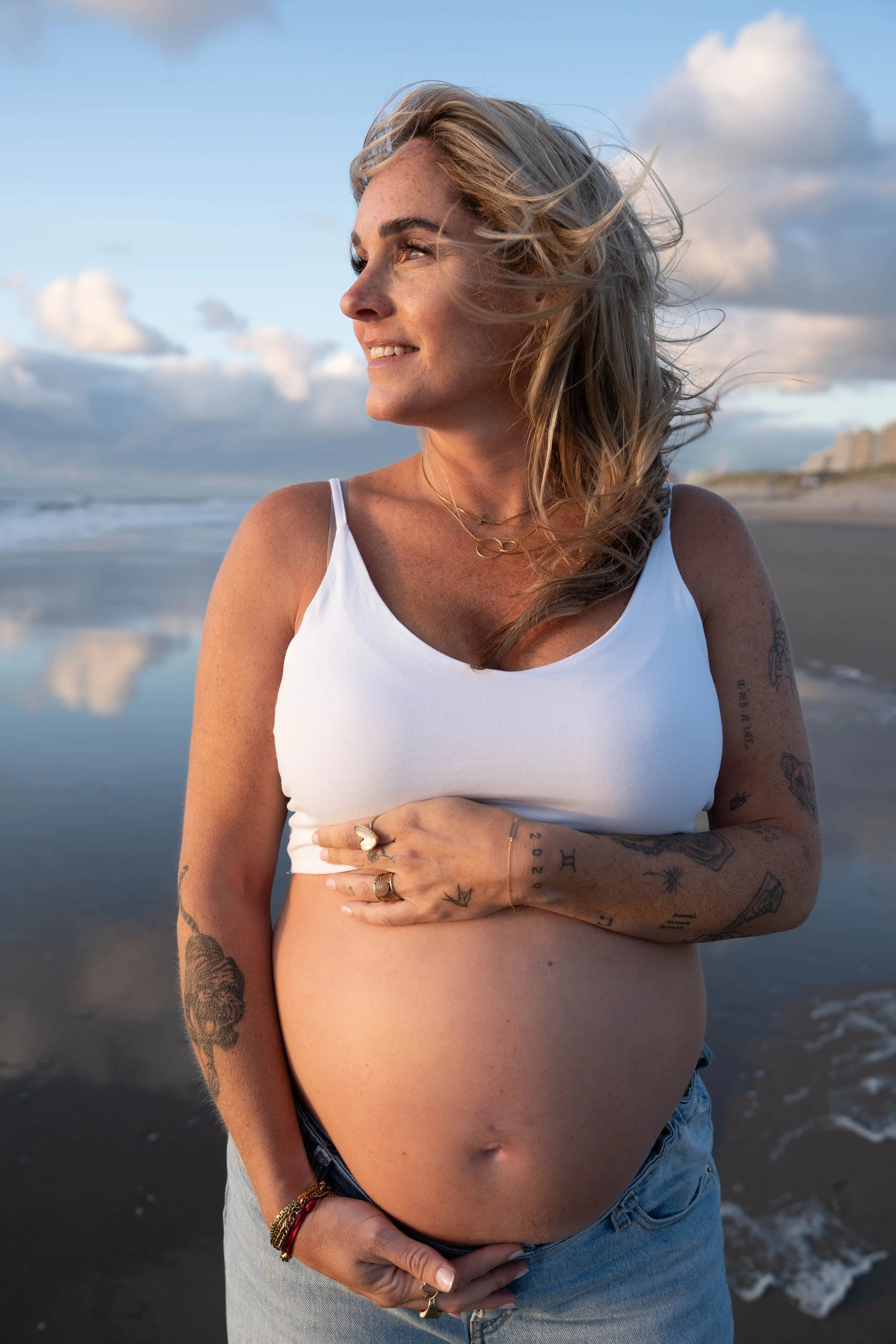 A woman with blonde hair standing on a beach, wearing a white crop top and jeans, with her hand on her pregnant belly, looking to the side with a smile. The background shows sand, water, and a partly cloudy sky.