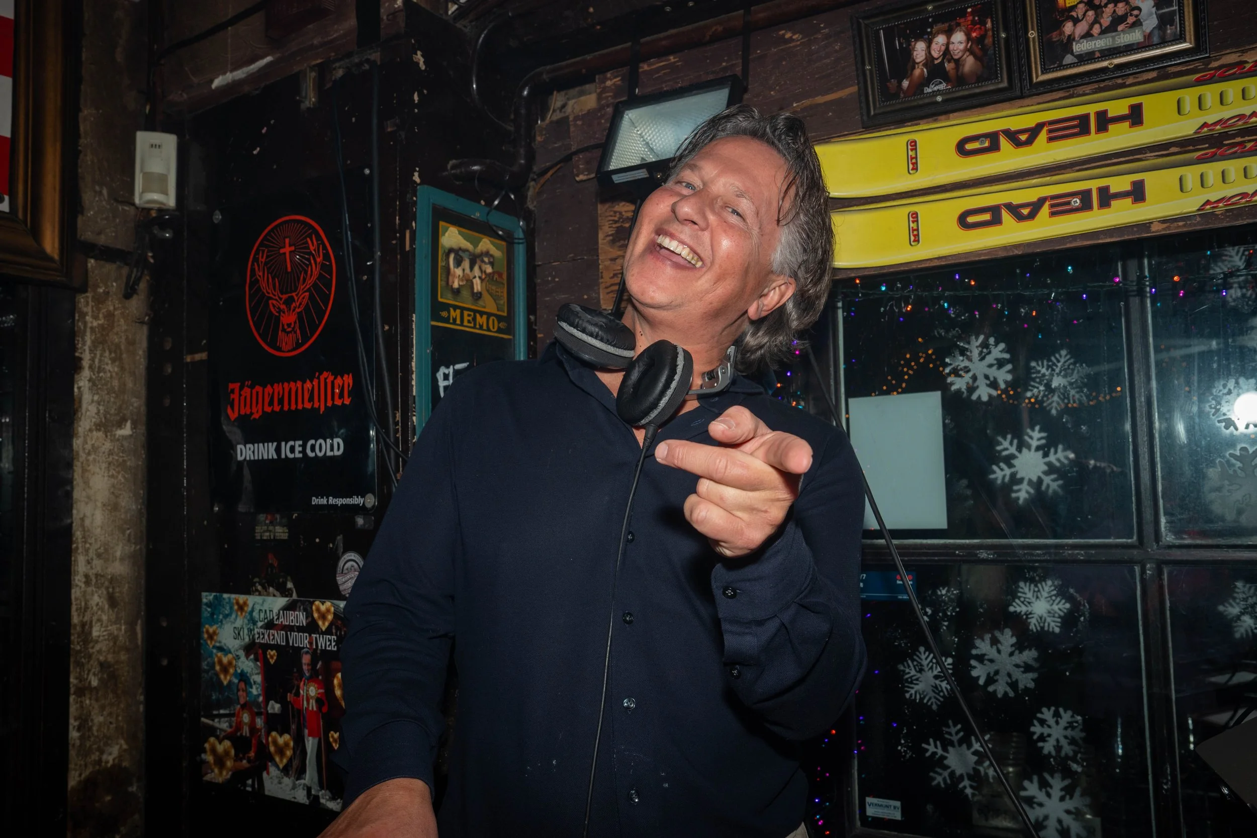 A man with gray hair smiling, wearing a dark shirt and a headset around his neck, standing in a bar or restaurant decorated with framed pictures, neon signs, and Christmas snowflake decorations.
