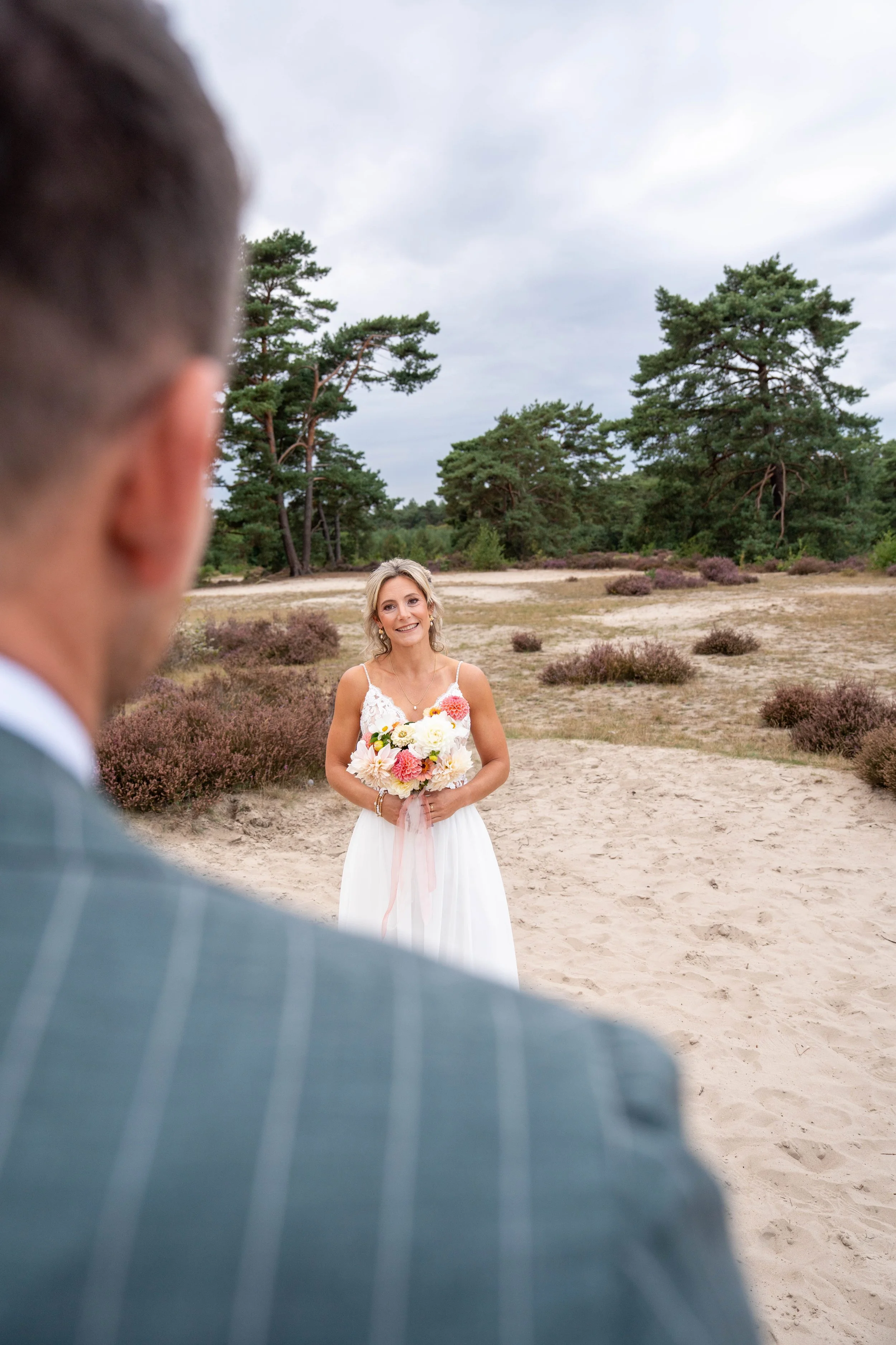 A bride holding a bouquet of flowers smiles at a groom during a wedding ceremony outdoors in a sandy area with purple bushes and tall green trees under a cloudy sky. The groom is partially visible in the foreground, out of focus.