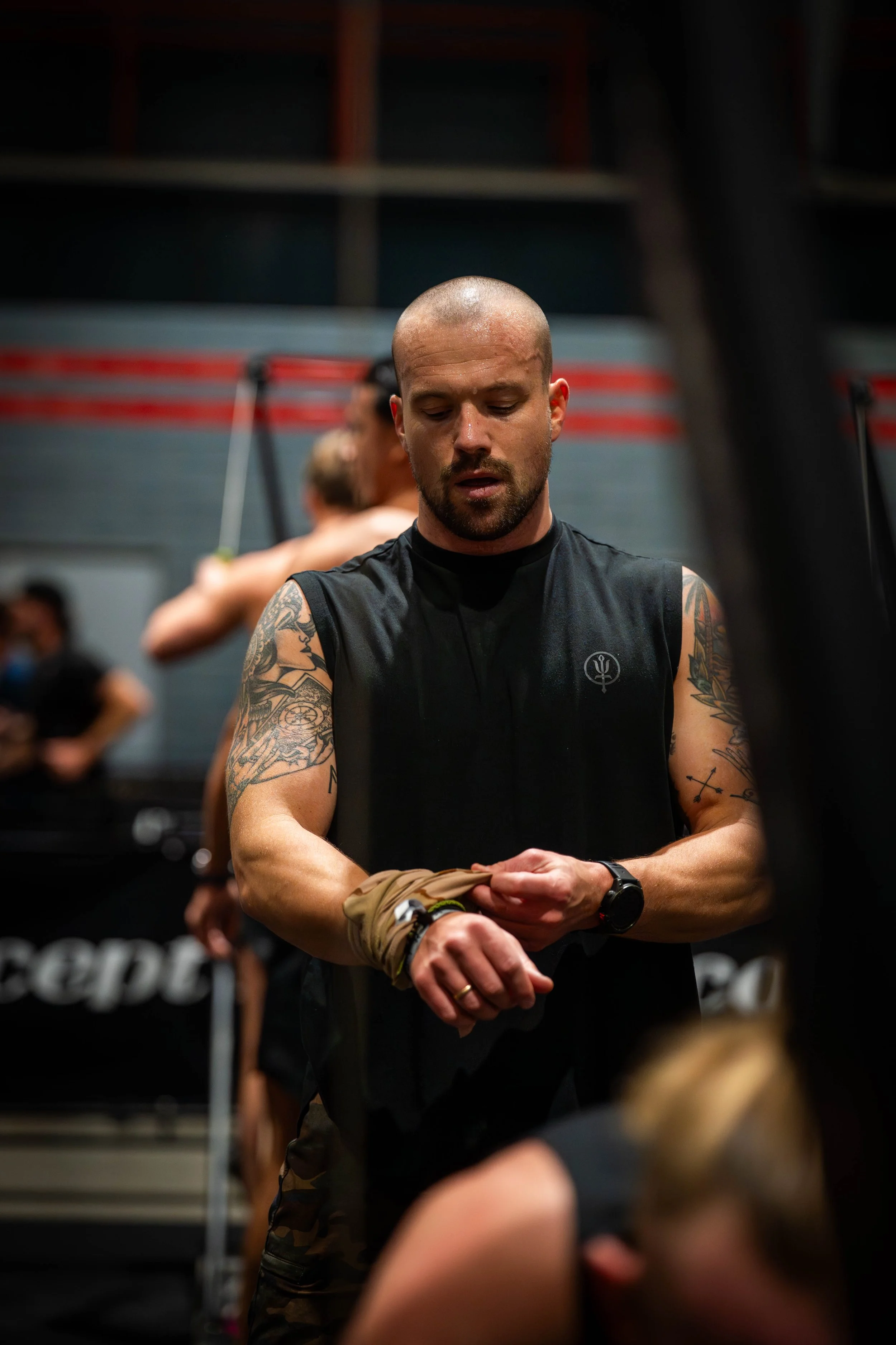 An individual with a shaved head and tattoos on their arms, wearing a black sleeveless shirt, standing in a gym or fitness facility with other people in the background.