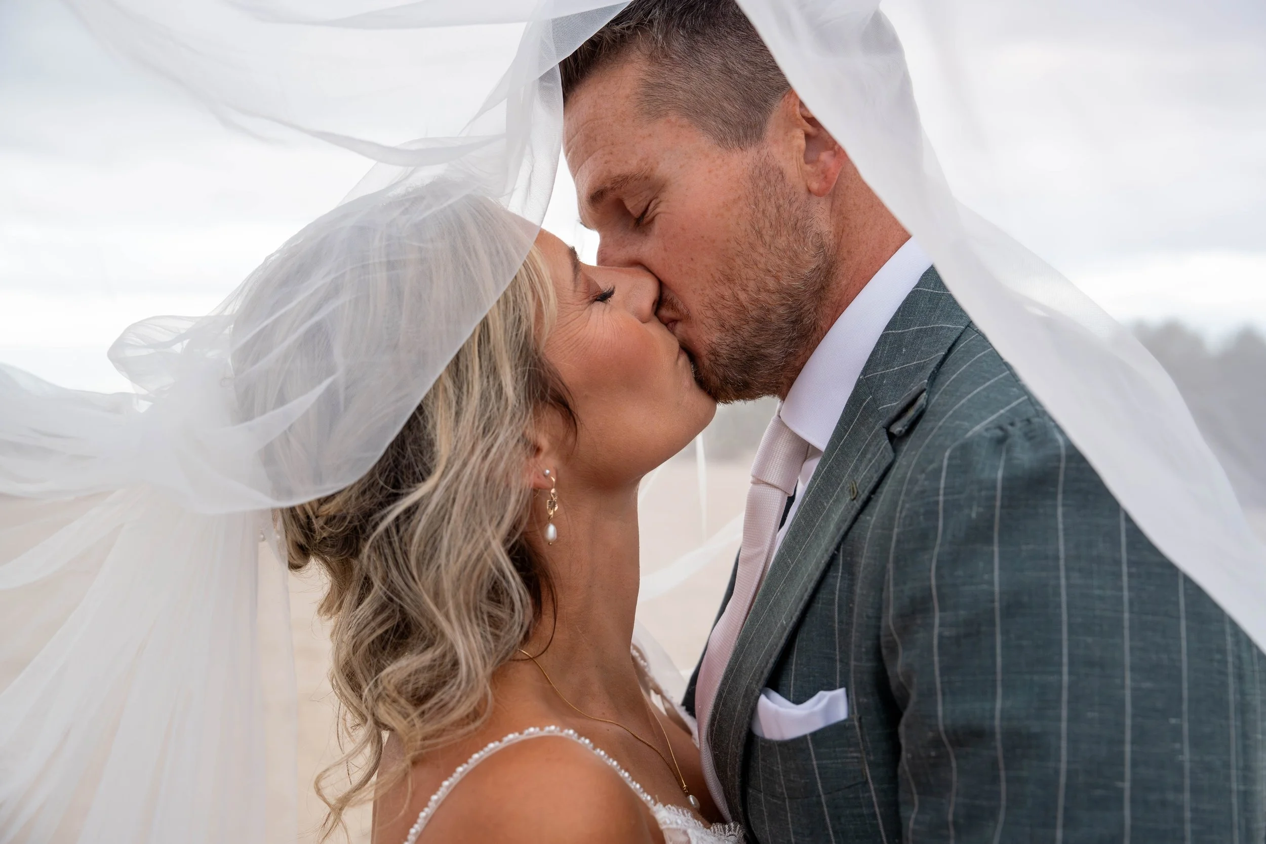 A bride and groom sharing a kiss under the bride's veil during their wedding. The groom is wearing a checkered suit and the bride has long, wavy blonde hair and is wearing pearl earrings.
