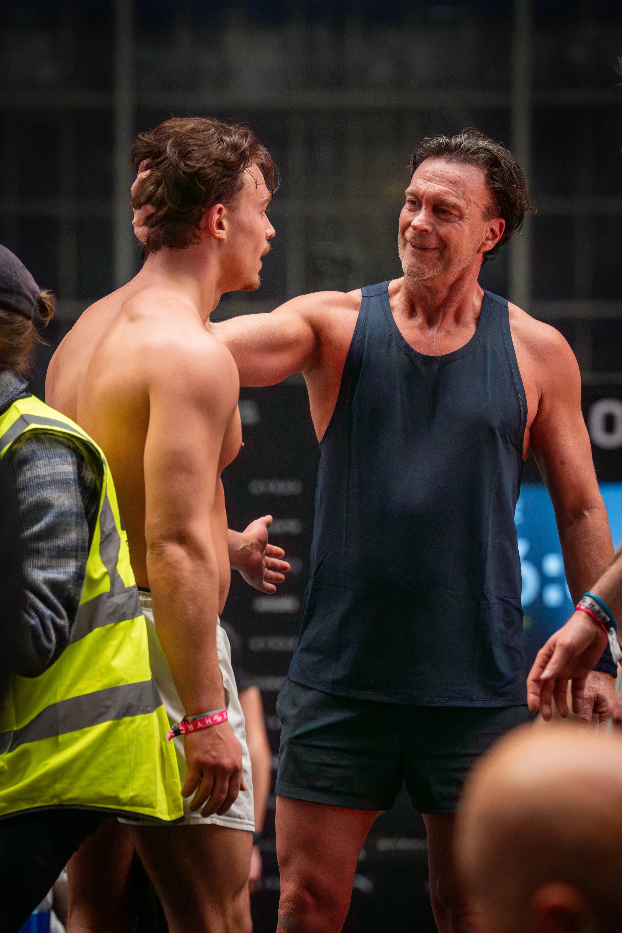 Two men, one shirtless and one in athletic wear, are having a conversation at what appears to be a sports event, with one man smiling and the other, shirtless man, looking serious. There are people around them and a dark background.