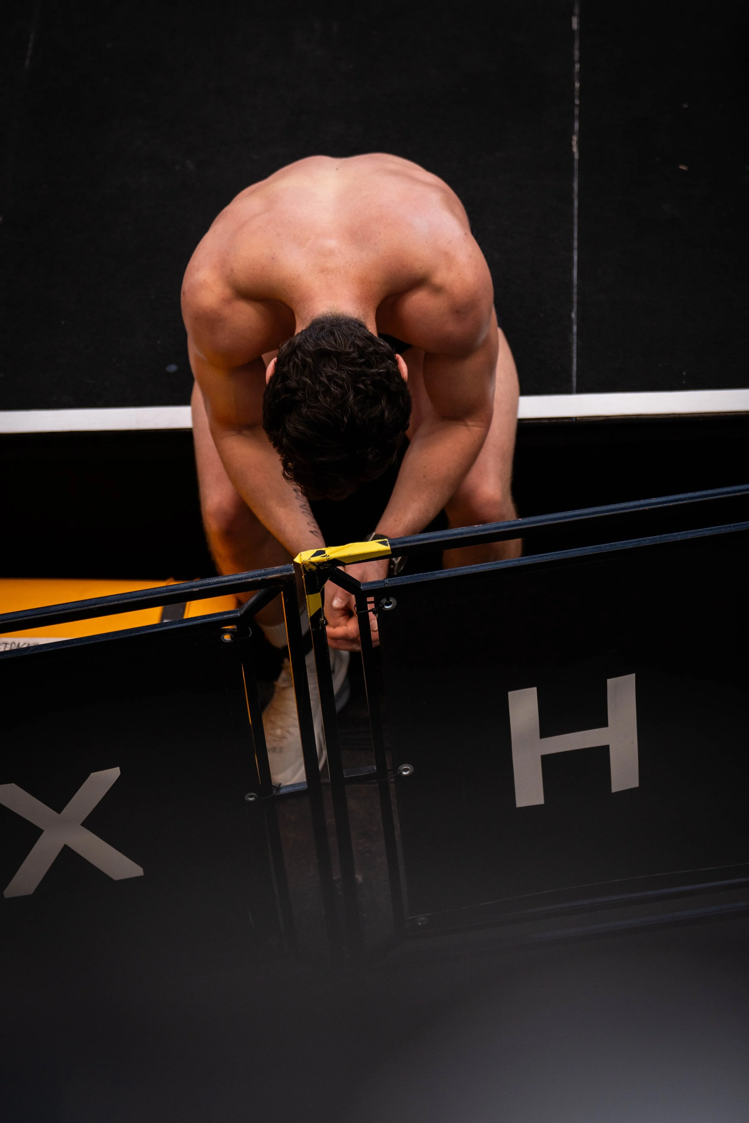 A person shirtless on a black stage, bent over with arms extended downward, possibly preparing for a performance or stretching, with stage markings and a railing in the foreground.
