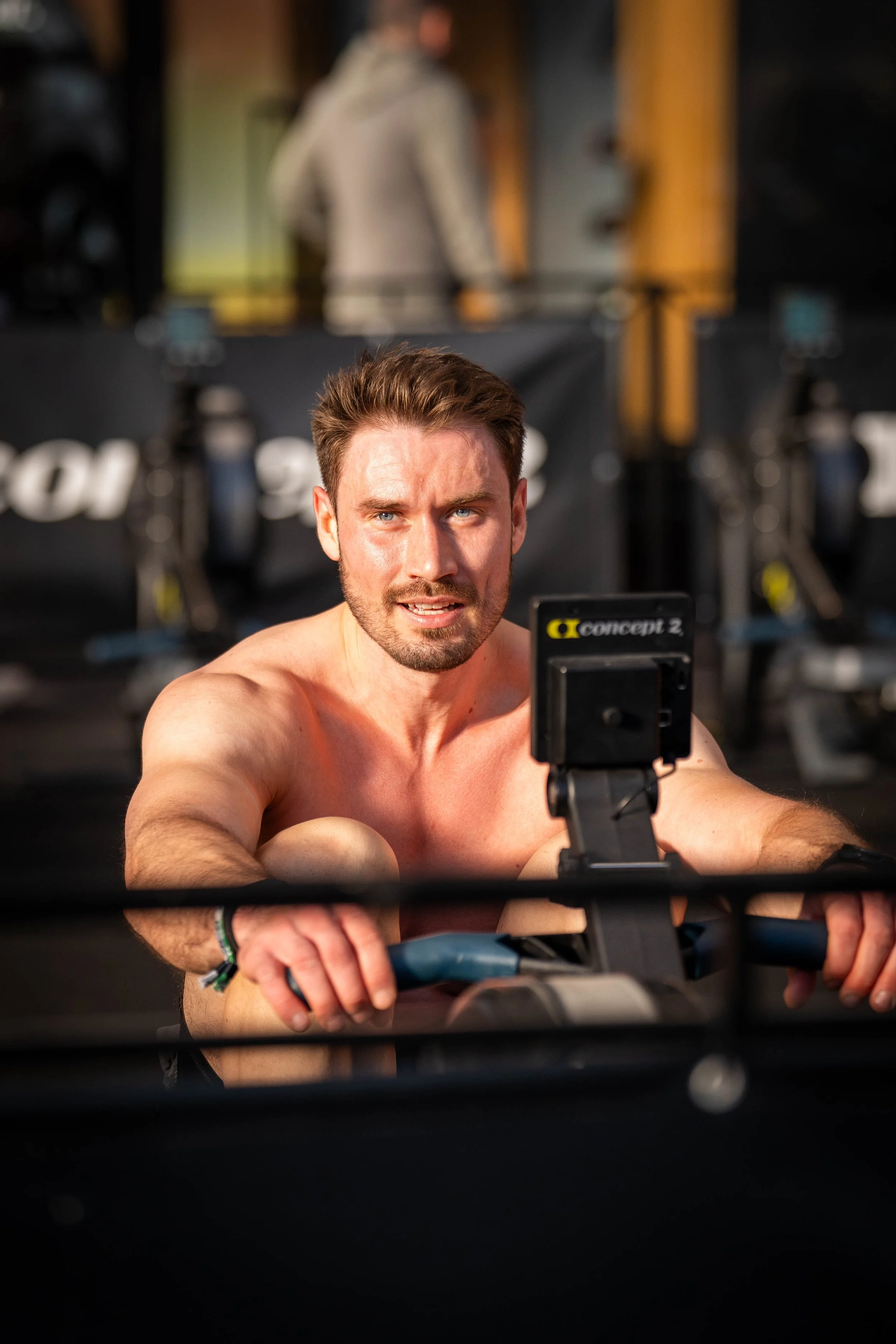 A shirtless man with short brown hair and blue eyes is exercising on a stationary bike in a gym, taking a selfie with his phone. In the background, a large mirror reflects a person in a beige hoodie, and gym equipment is visible.