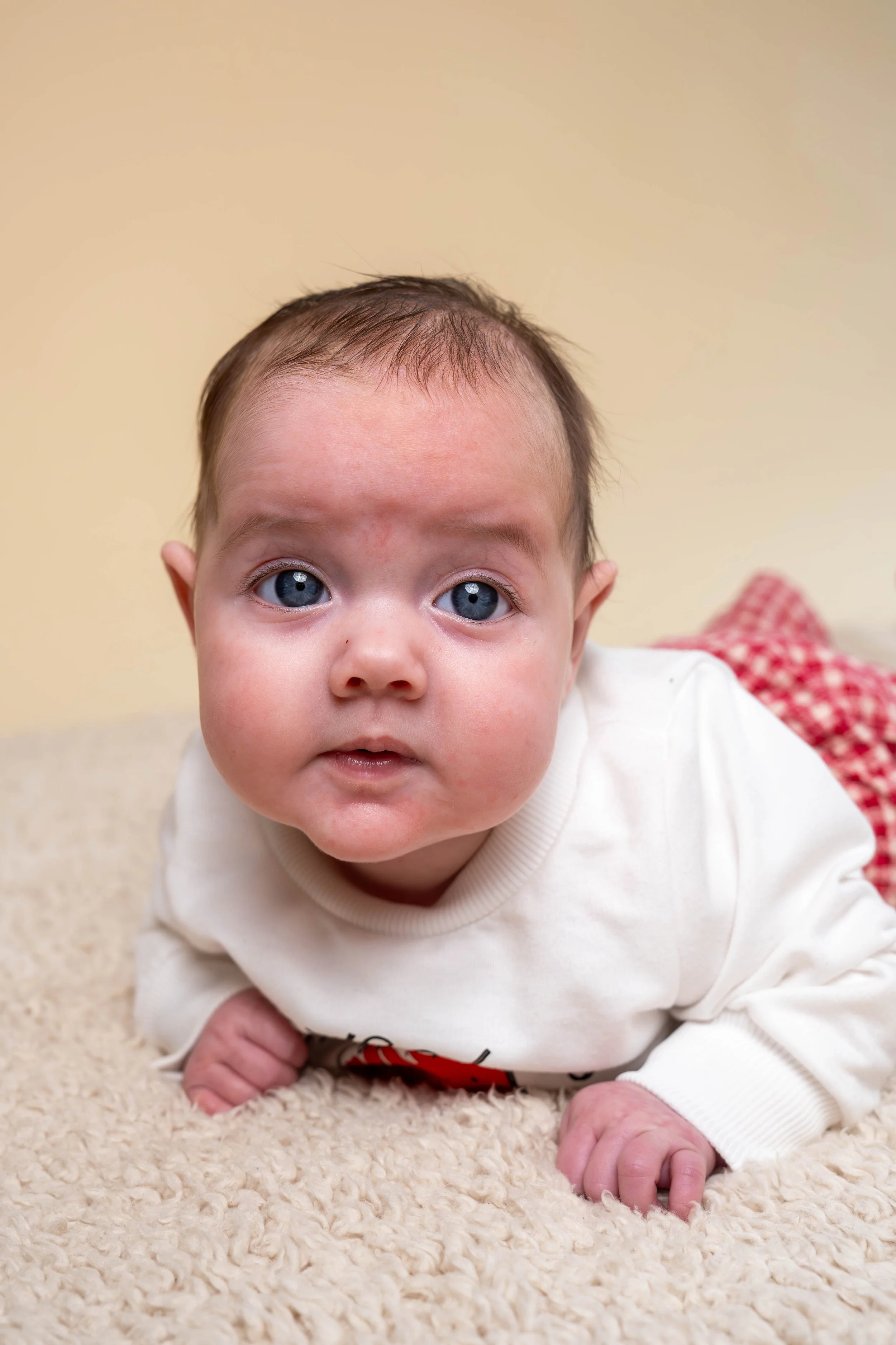 Close-up of a baby crawling on a soft, beige carpet, with light skin, blue eyes, and dark hair, wearing a cream-colored long-sleeve shirt.