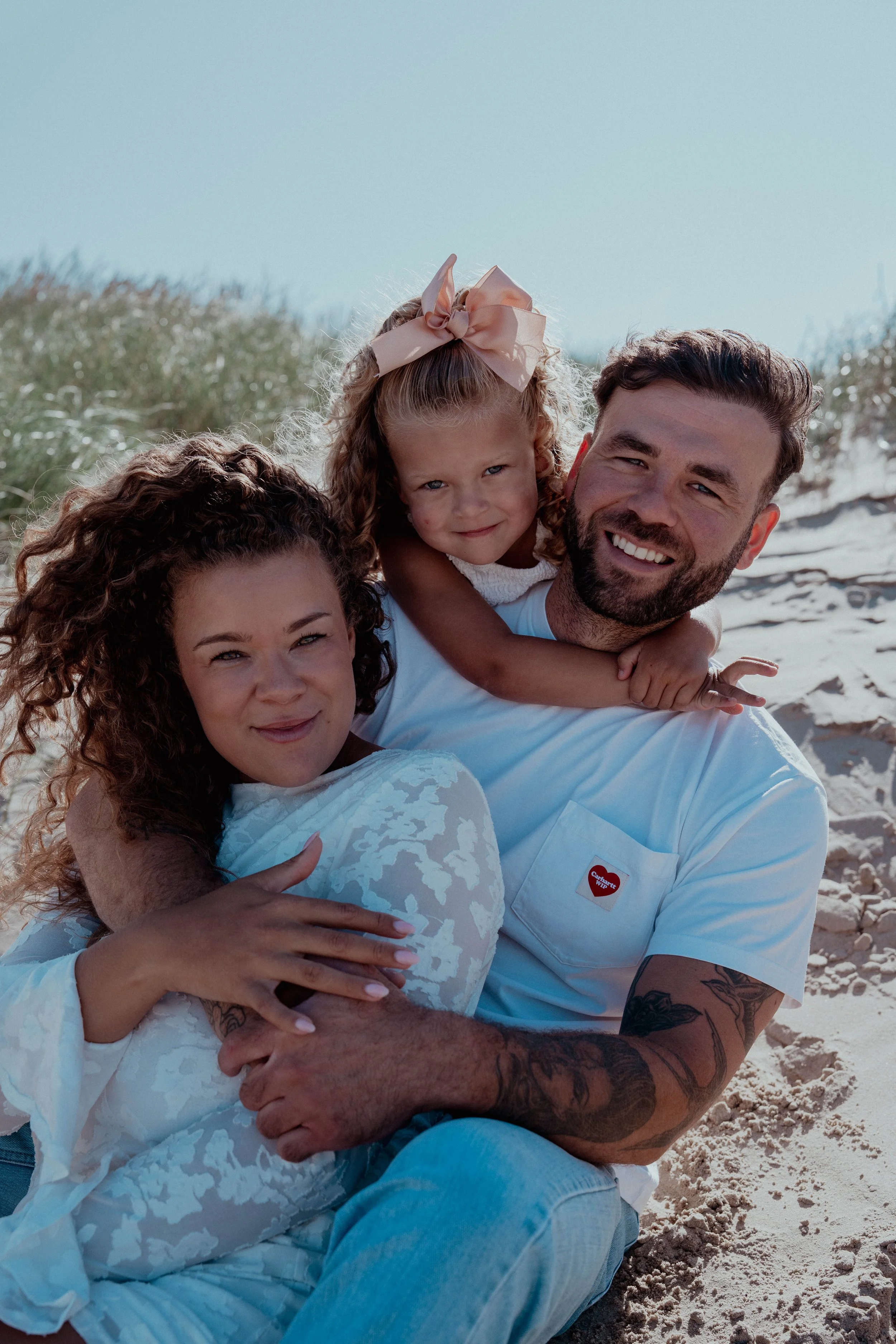 A happy family at the beach, with three people - a woman with curly hair, a man with a beard, and a little girl with a pink bow in her hair. They are smiling and cuddling.