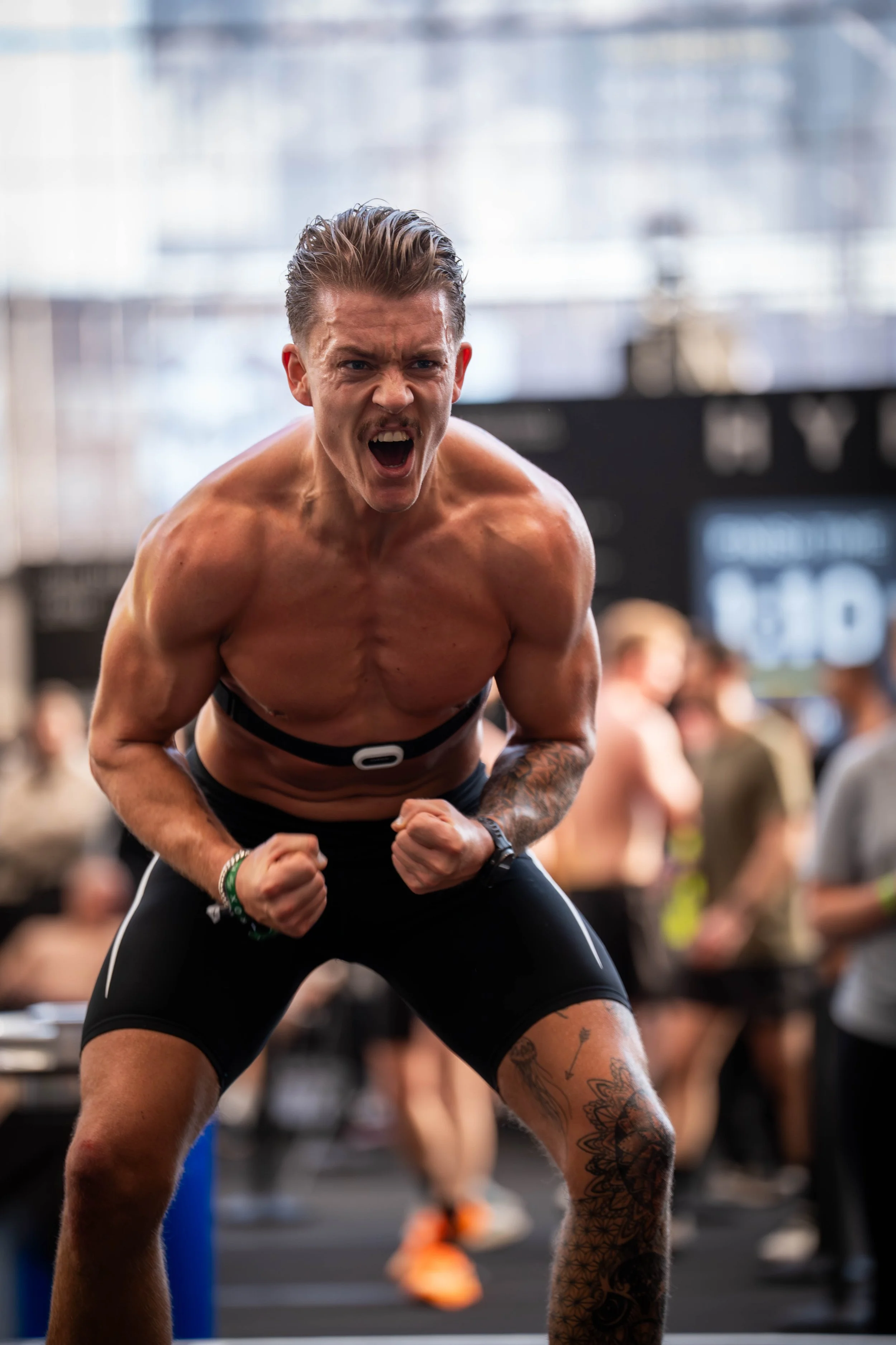A muscular man with tattoos on his left leg and a tattoo on his right forearm is shouting with a fierce expression, clenching his fists, and leaning forward in a workout gym with a group of people in the background.