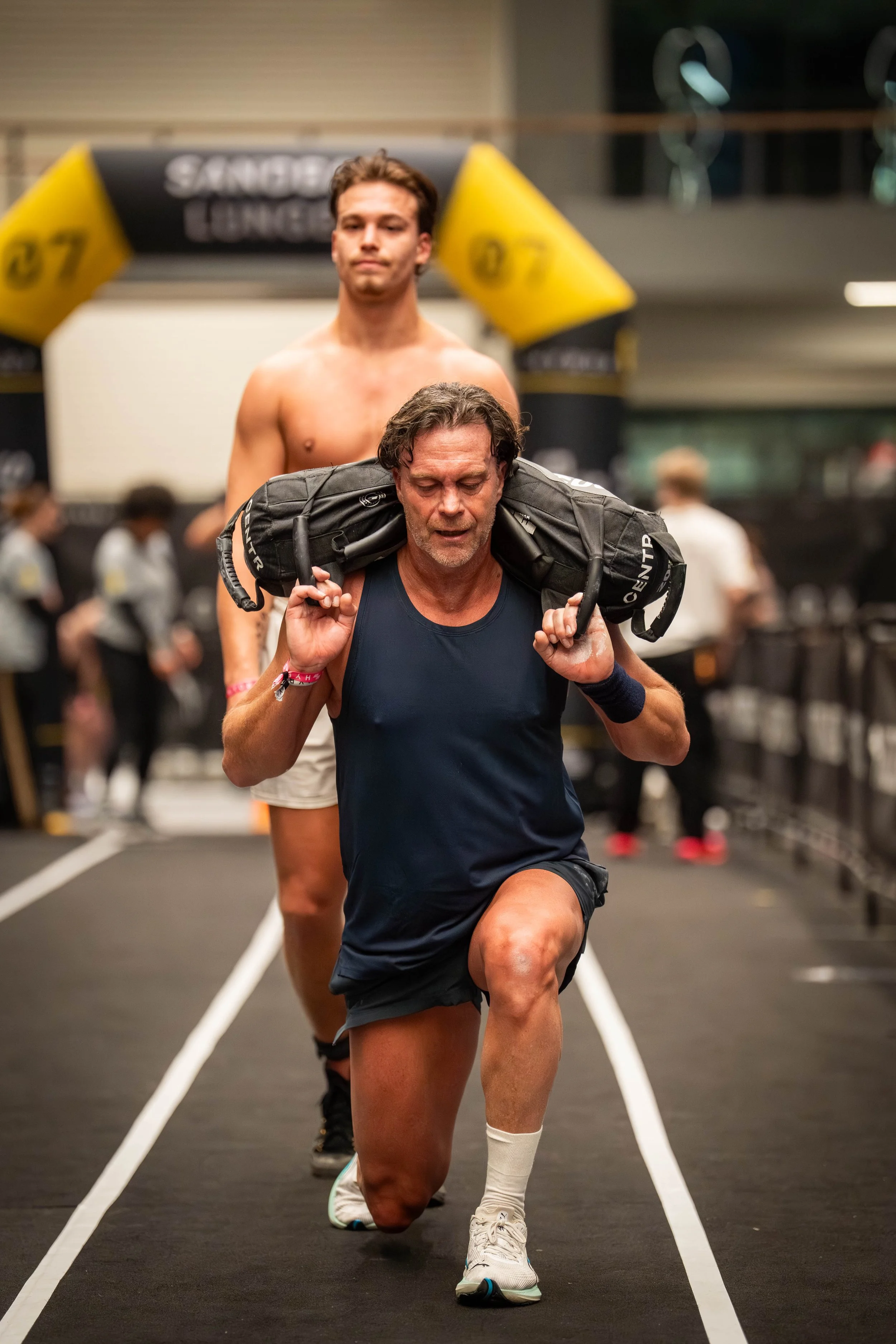 HYROX fotografie van een atleet tijdens het sandbag carry onderdeel in een indoor wedstrijdomgeving.