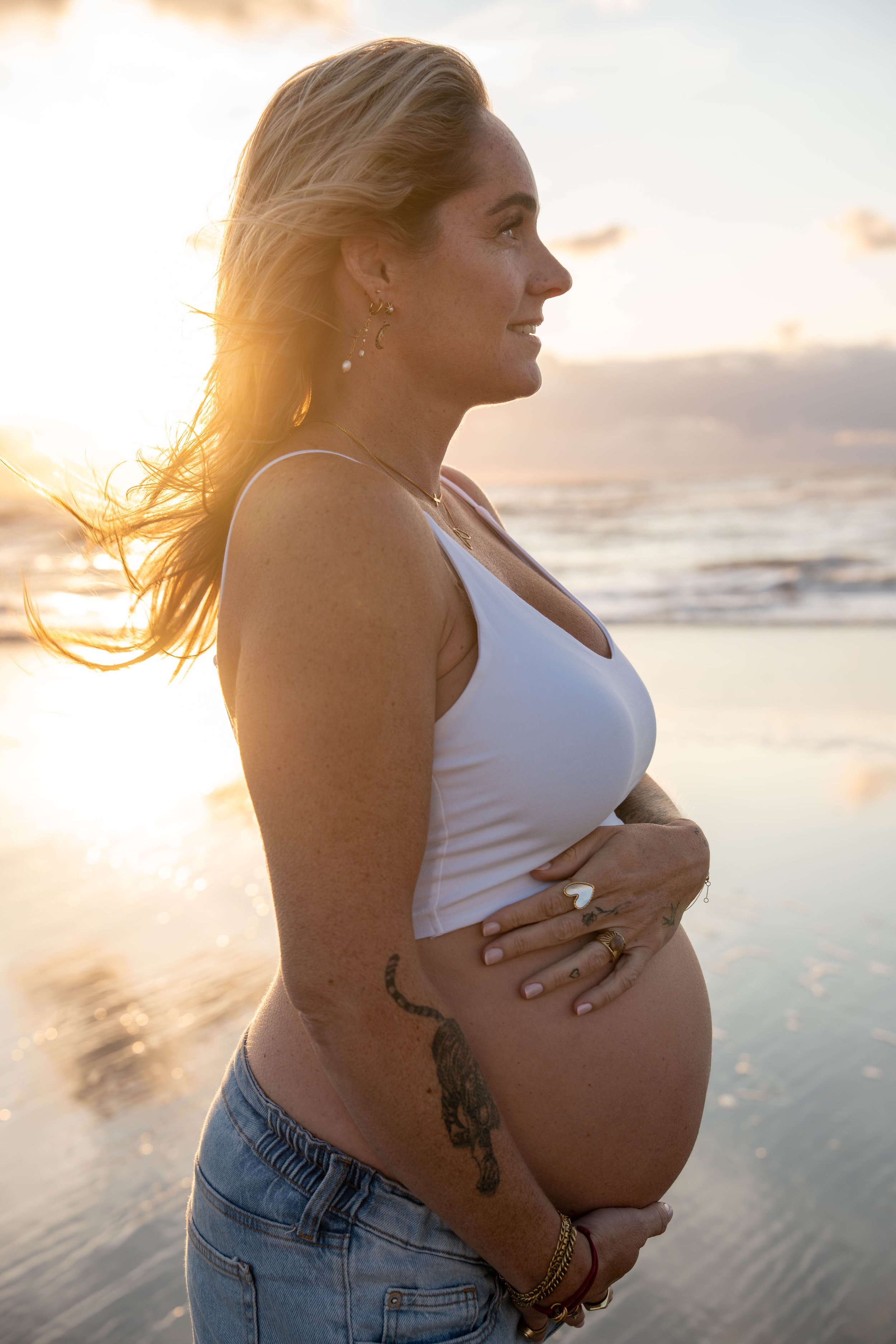 Pregnant woman in a white tank top standing on the beach at sunset, looking to her right with her hands resting on her belly, wearing earrings and jewelry, with ocean waves in the background.