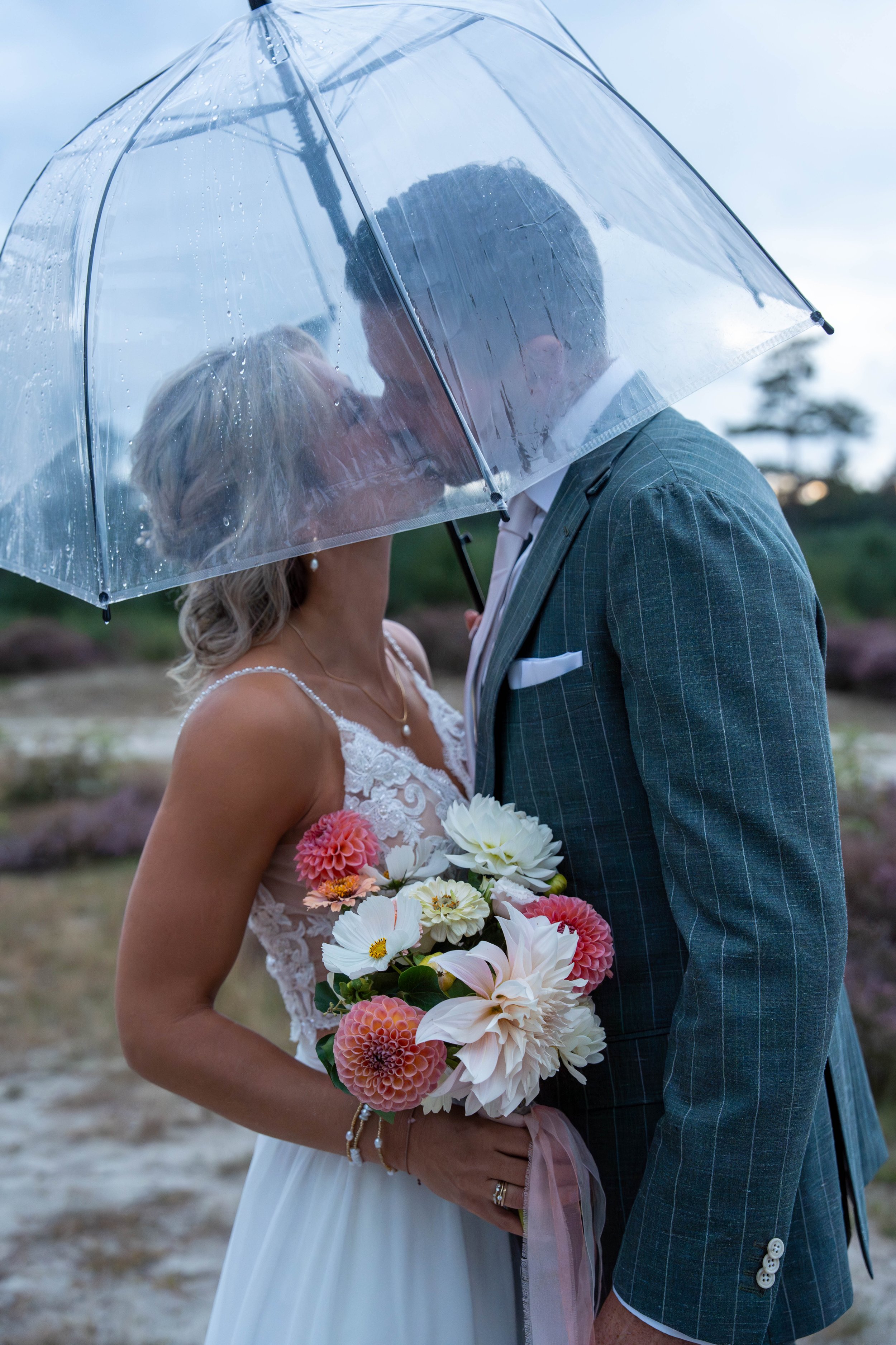 A bride and groom kiss under a clear umbrella, with the bride holding a bouquet of white and pink flowers, standing outdoors on a cloudy day.