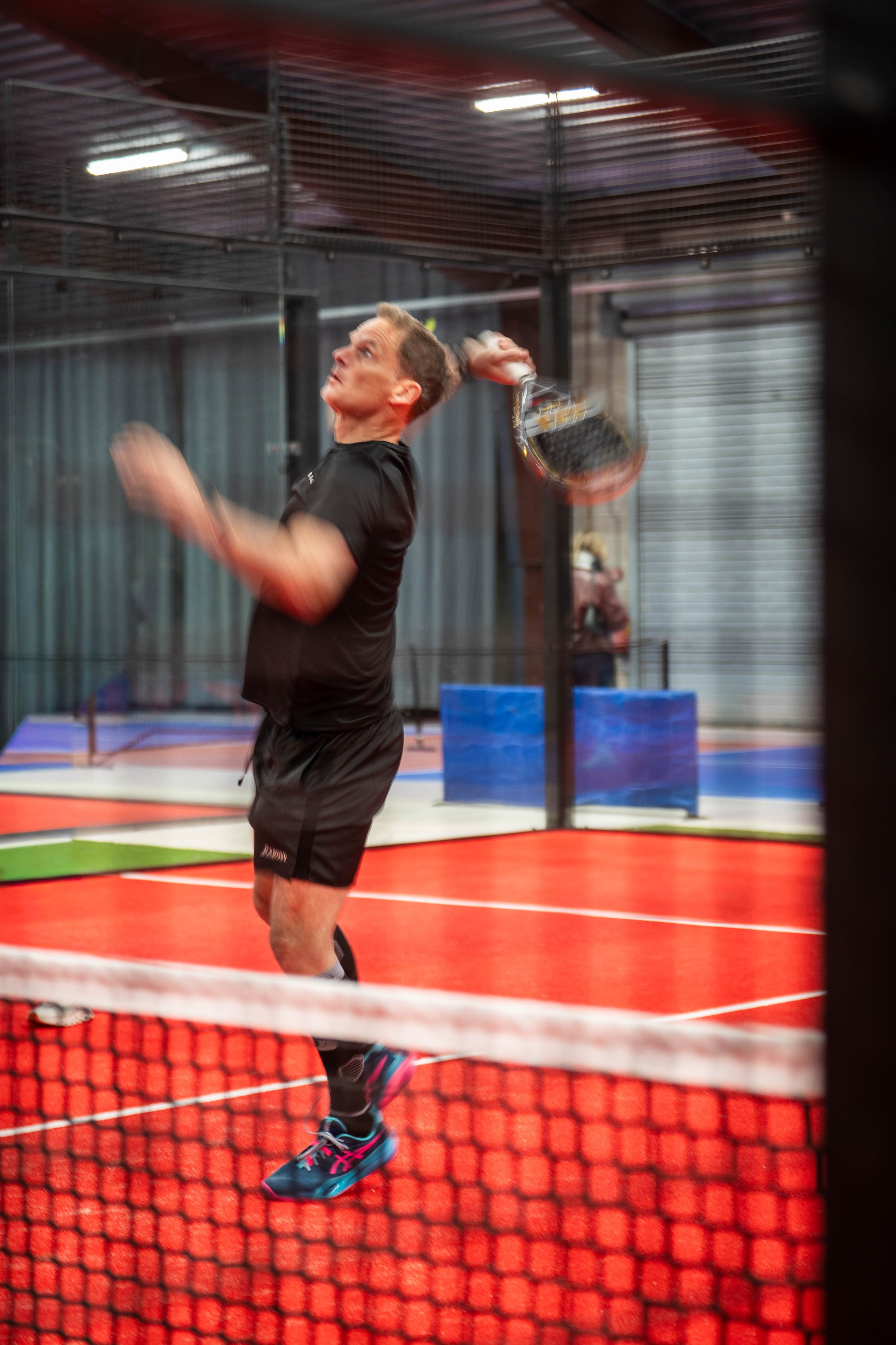A man playing tennis on an indoor court, seen through a net, with a focus on his side profile as he swings with a racket.