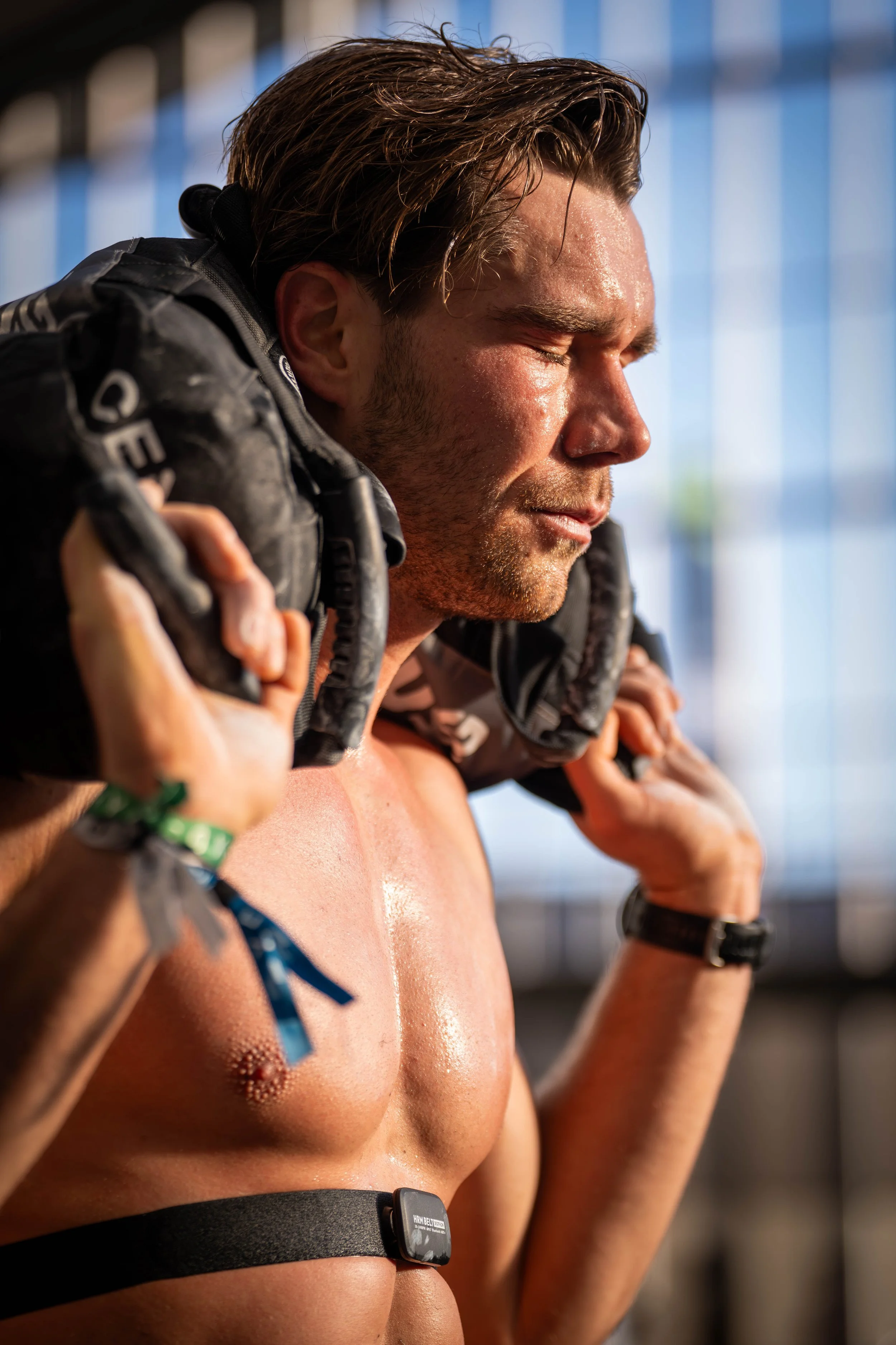 A man with wet skin, likely from exercise, is putting on a black jacket over his shoulders. He is shirtless, wearing a black strap with a device around his waist, and has a focused expression. The background features a blurred blue structure.