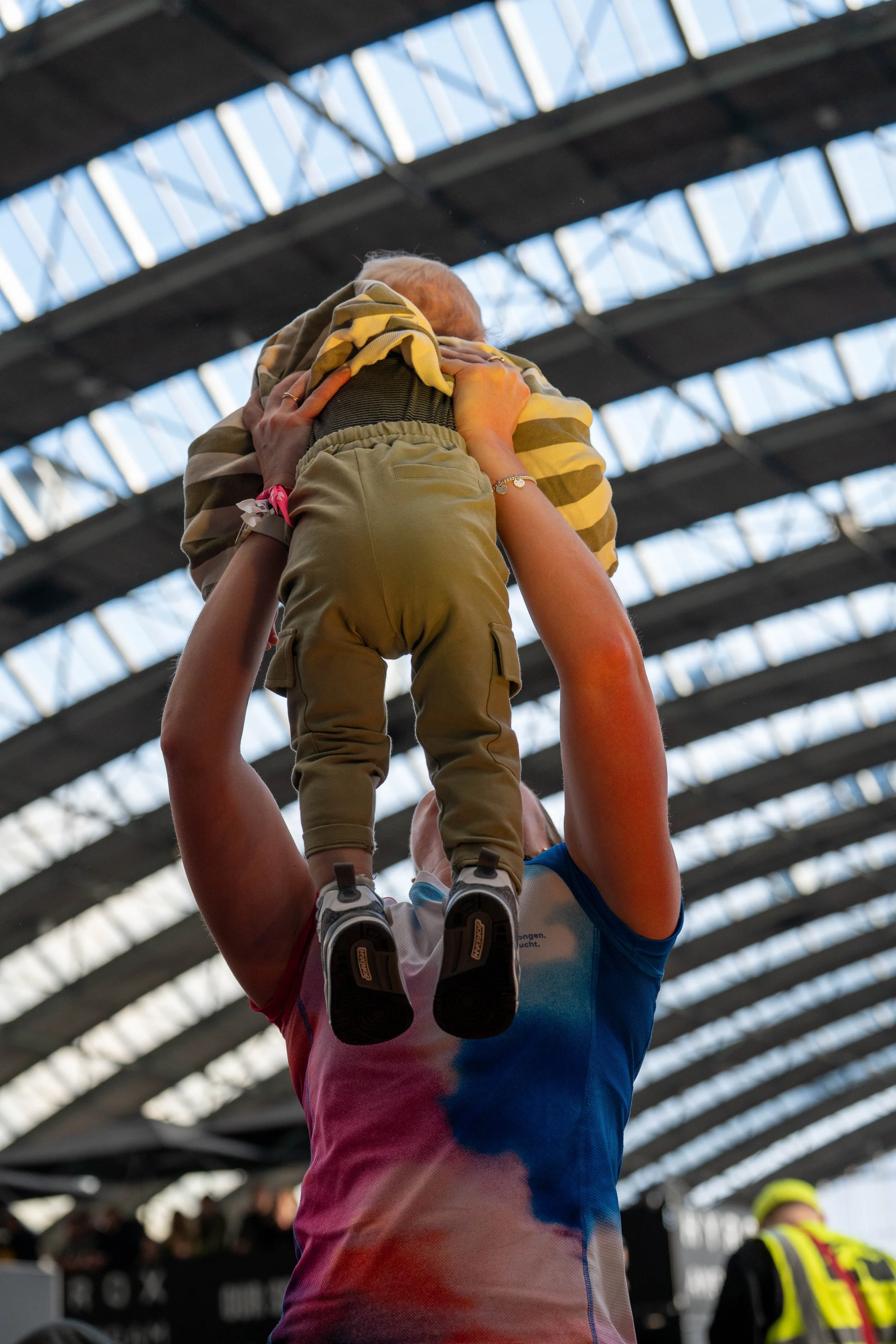 Person lifting a small child or baby in a stadium with a high ceiling and sunlight streaming through the broken roof panels.