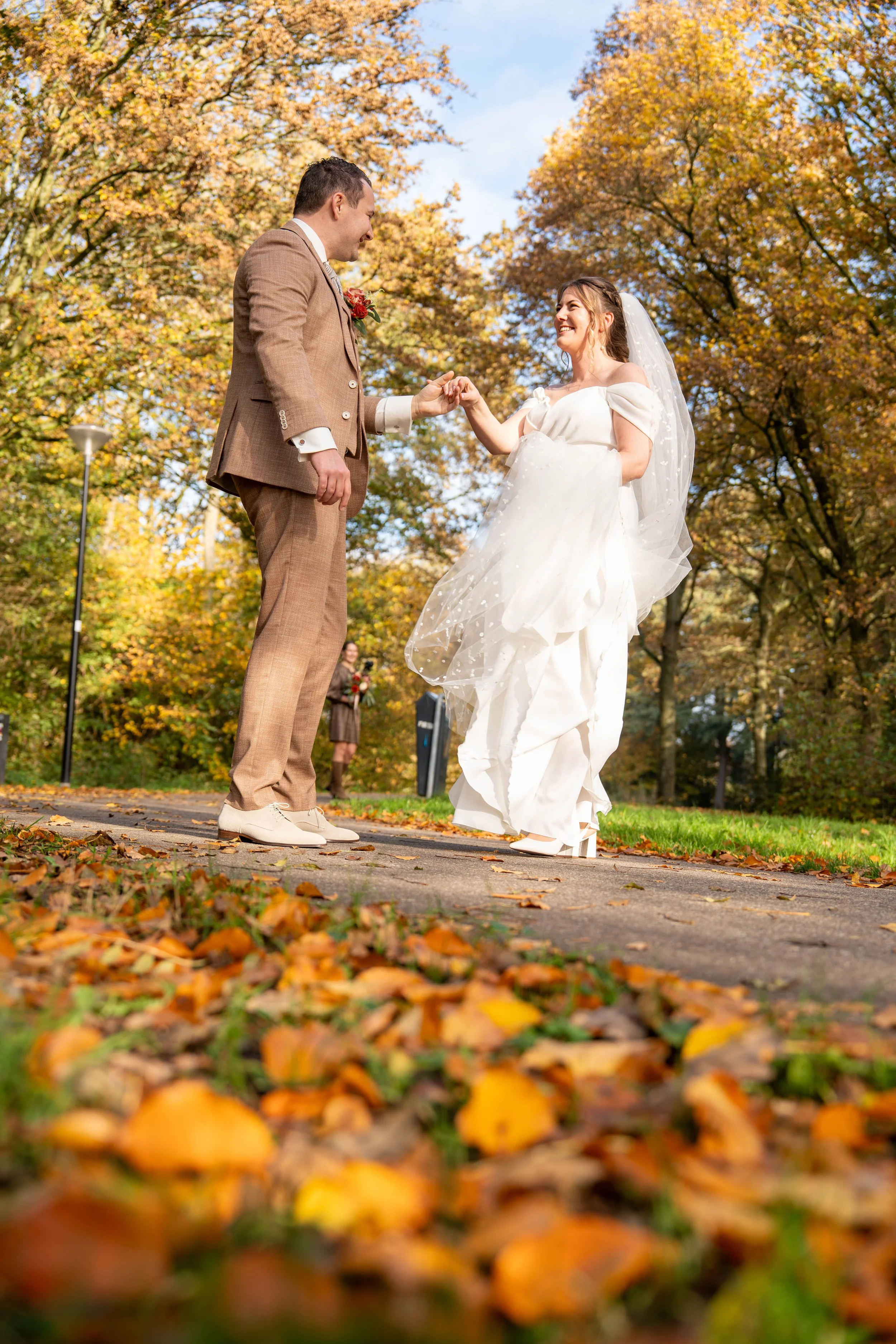 A bride and groom holding hands and smiling at each other outdoors during autumn, with fall foliage in the background.