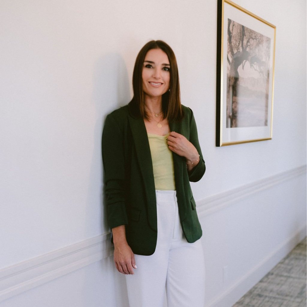A woman with shoulder-length brown hair and a smile, standing against a white wall with a framed picture of a tree in the background.