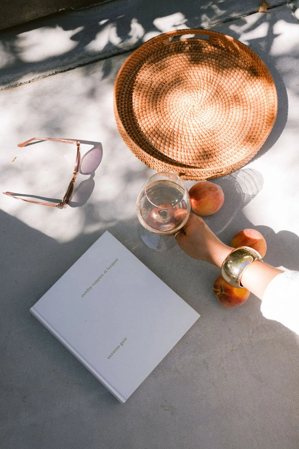 A book titled 'Sunday Suppers at Lucques' by Suzanne Goin lying on a gray surface, surrounded by four peaches, a glass of white wine, a pink pair of sunglasses, and a woven round basket. Shadows cast from nearby foliage create patterns on the surface.