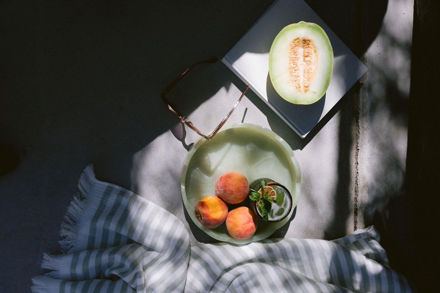 A still life scene with three peaches and a small plant on a green plate, a sliced melon on a gray plate, a pair of sunglasses, a striped cloth, and a white surface, with sunlight casting shadows.