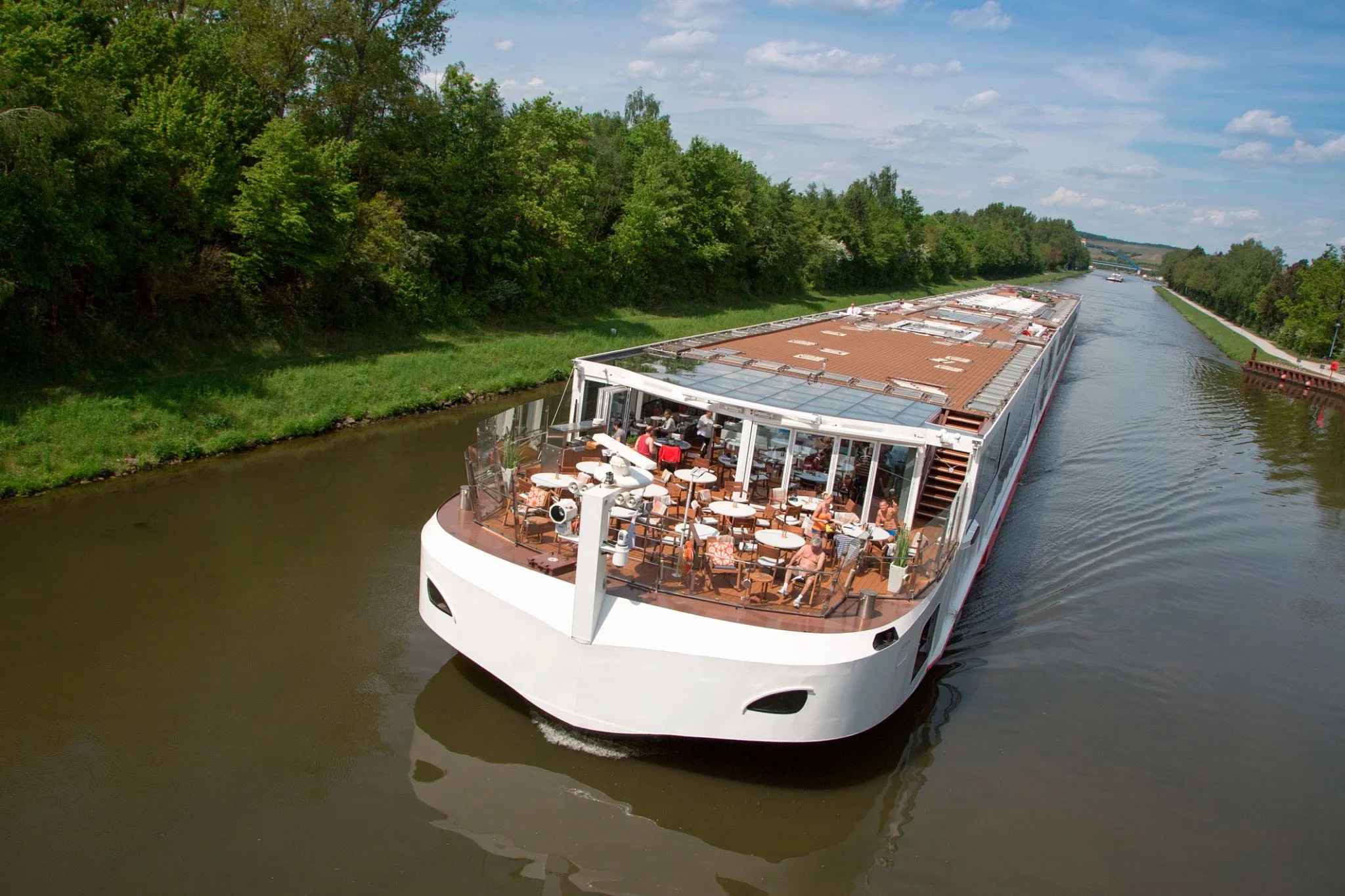 A large white cruise boat with an outdoor deck filled with people, sailing along a calm river surrounded by green trees on a sunny day.