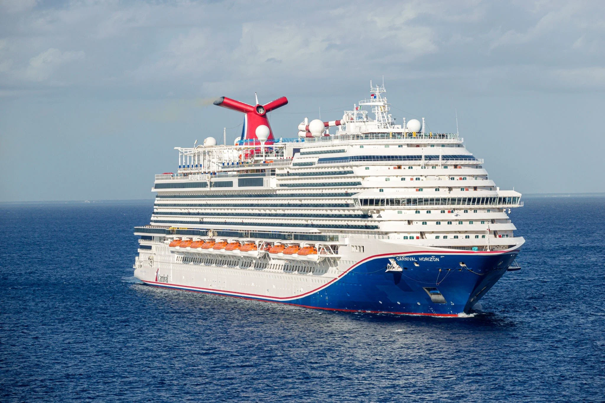 A large white cruise ship with multiple decks, orange lifeboats, and a red and black funnel sailing on the ocean under a cloudy sky.
