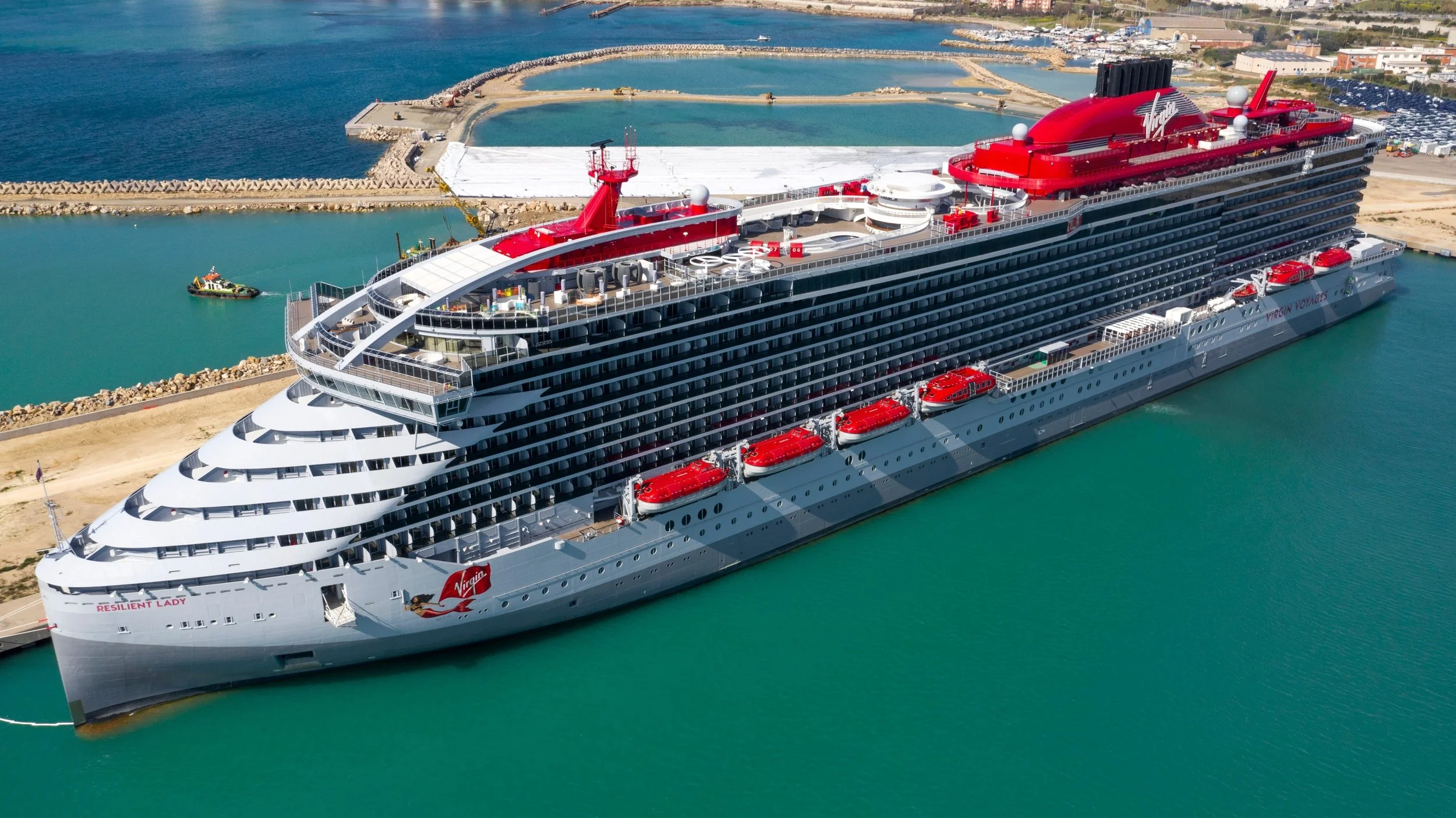 A large cruise ship docked at a port with calm water, multiple decks, amenities on top, and a red funnel with the Virgin logo.