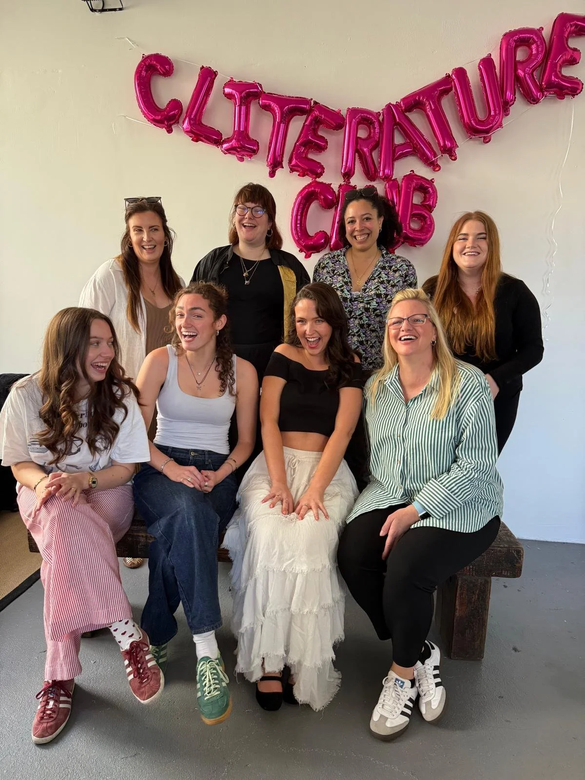 Group of nine women smiling and sitting together in front of pink foil balloon letters spelling 'Cliterature Club' on a white wall.