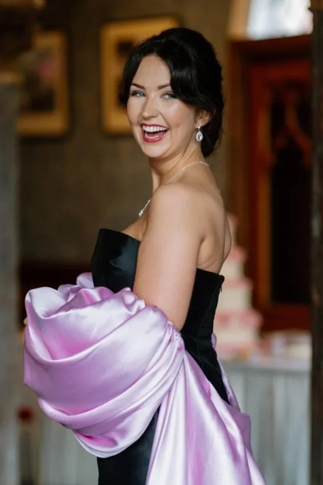 A young woman in a strapless black dress with pink satin fabric draped over her arm, smiling and looking over her shoulder in an indoor setting with framed pictures on the wall.