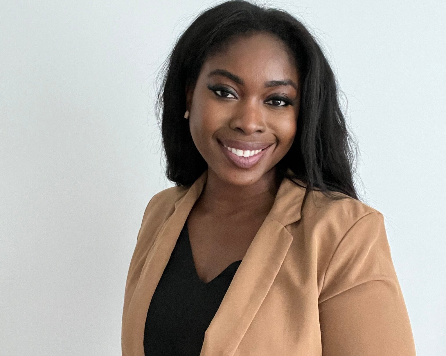 A smiling woman with black hair, wearing a tan blazer and black top, standing against a plain white background.
