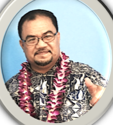 Man wearing glasses and a patterned shirt, with a purple lei around his neck, smiling indoors against a light blue background.