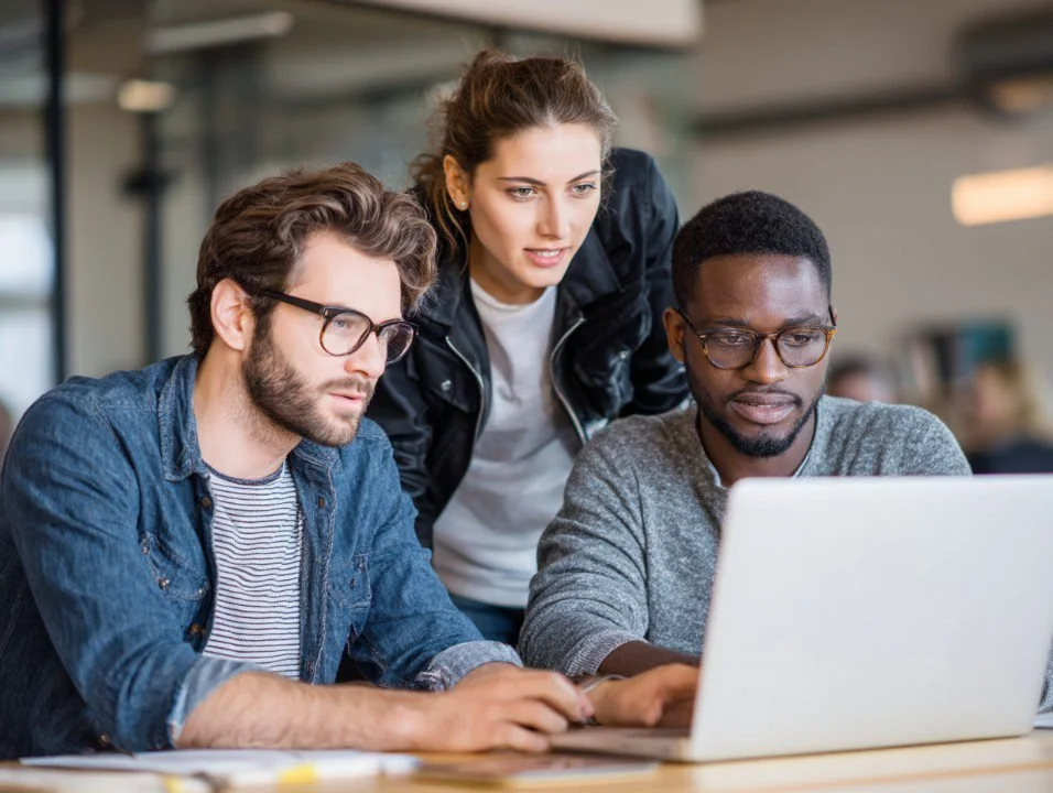 Three young adults working together on a laptop in an office setting