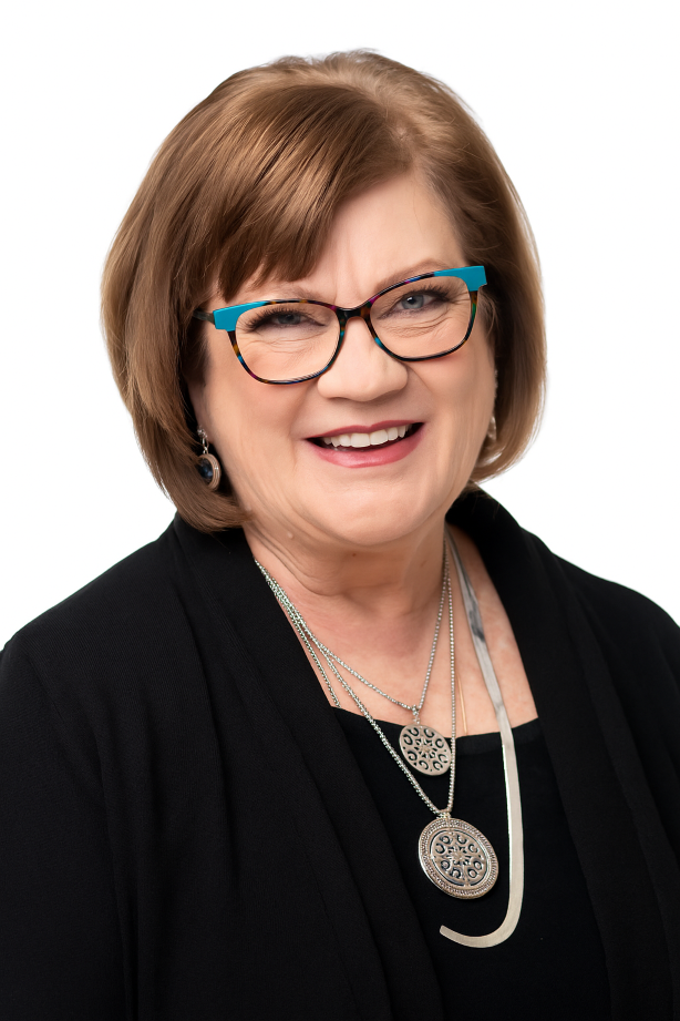 A smiling woman with short brown hair, wearing glasses with blue frames, earrings, and layered necklaces, dressed in a black blazer against a white background.