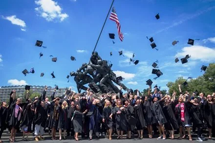 Group of graduates in caps and gowns celebrating under a sculpture on graduation day.