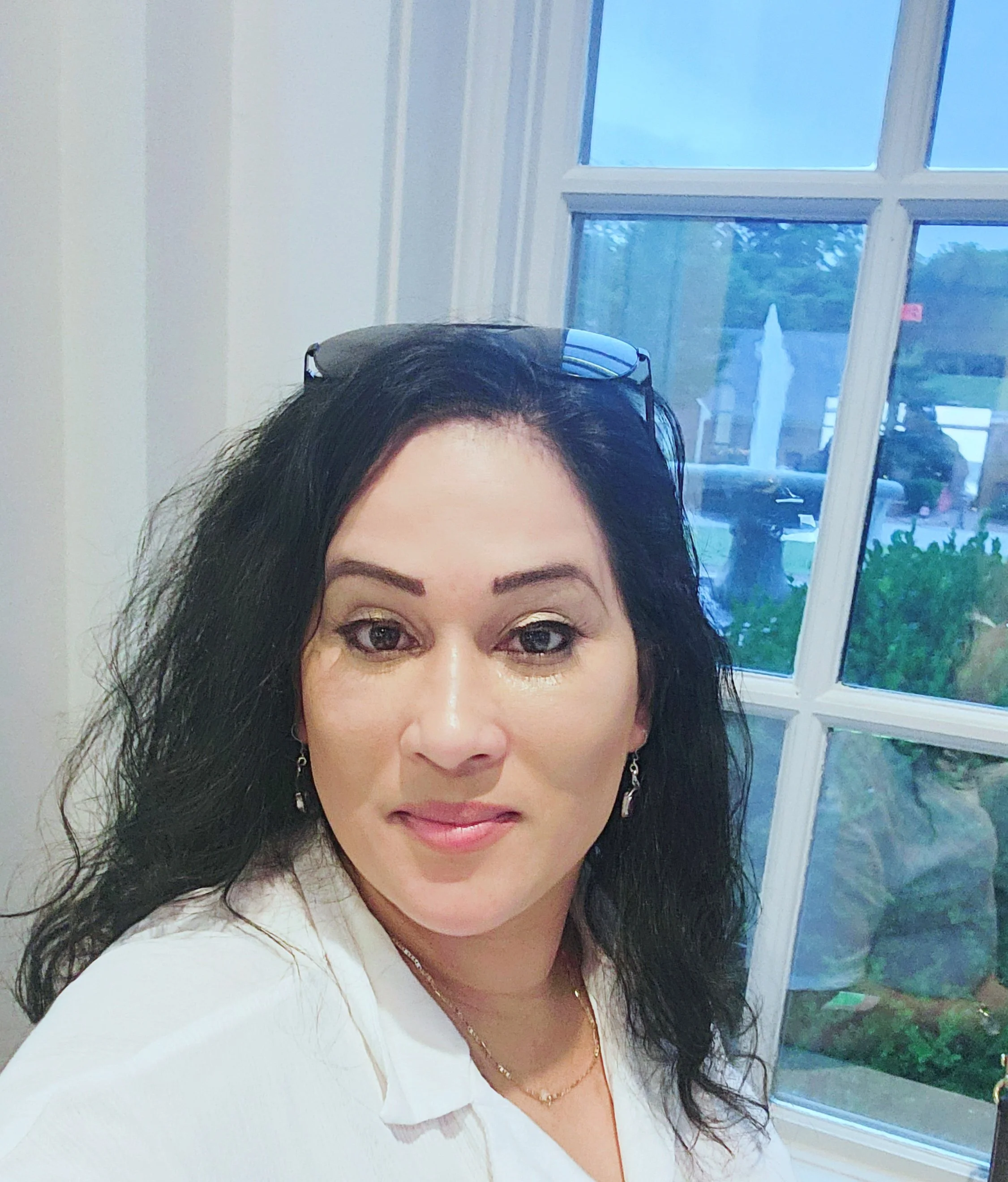 A woman with dark hair, wearing a white top and earrings, sitting near a window with a view of a parking lot and trees outside.