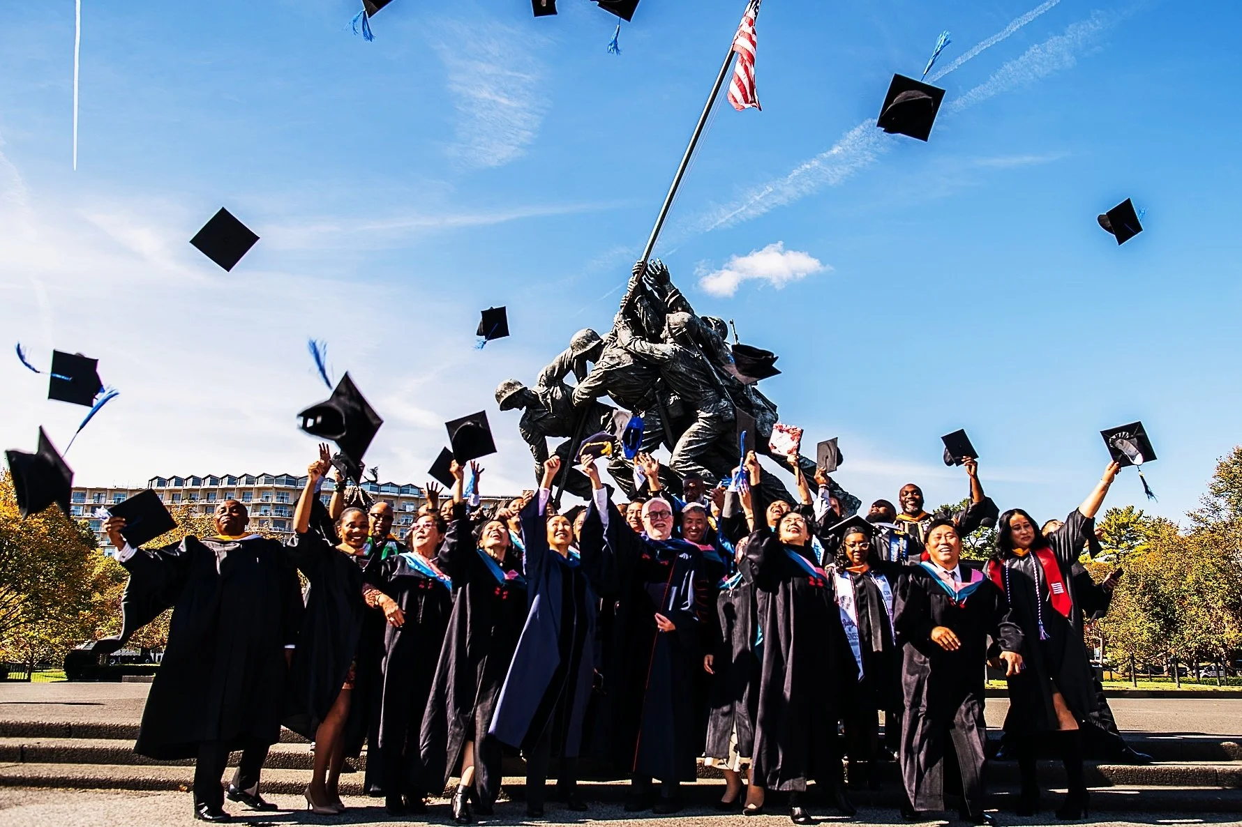 Graduates in caps and gowns throwing caps into the air during a graduation ceremony outdoors with the U.S. Marine Corps War Memorial in the background on a sunny day.